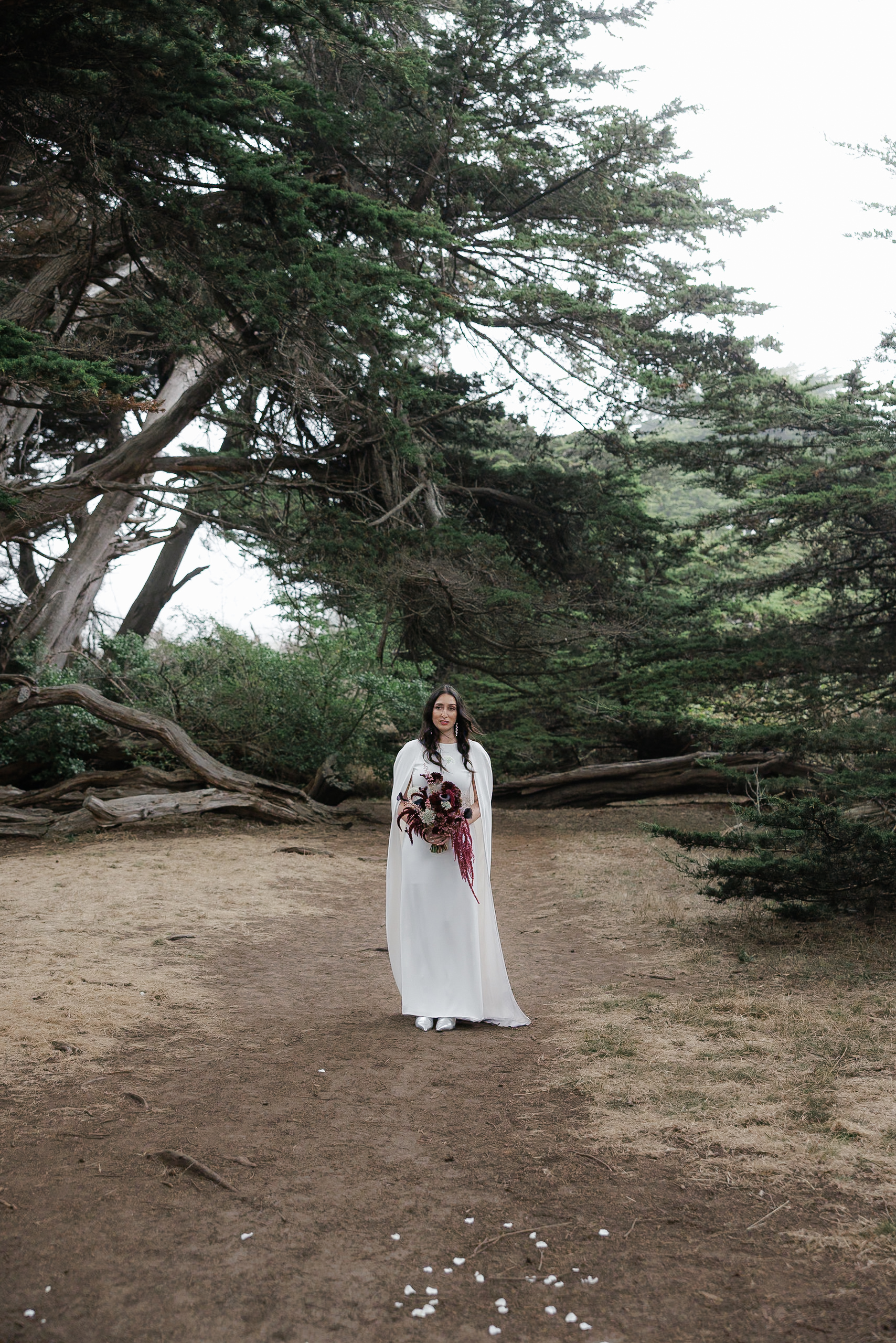 Mendocino forest wedding: Bride in white dress with bouquet on dirt path, surrounded by tall evergreens.