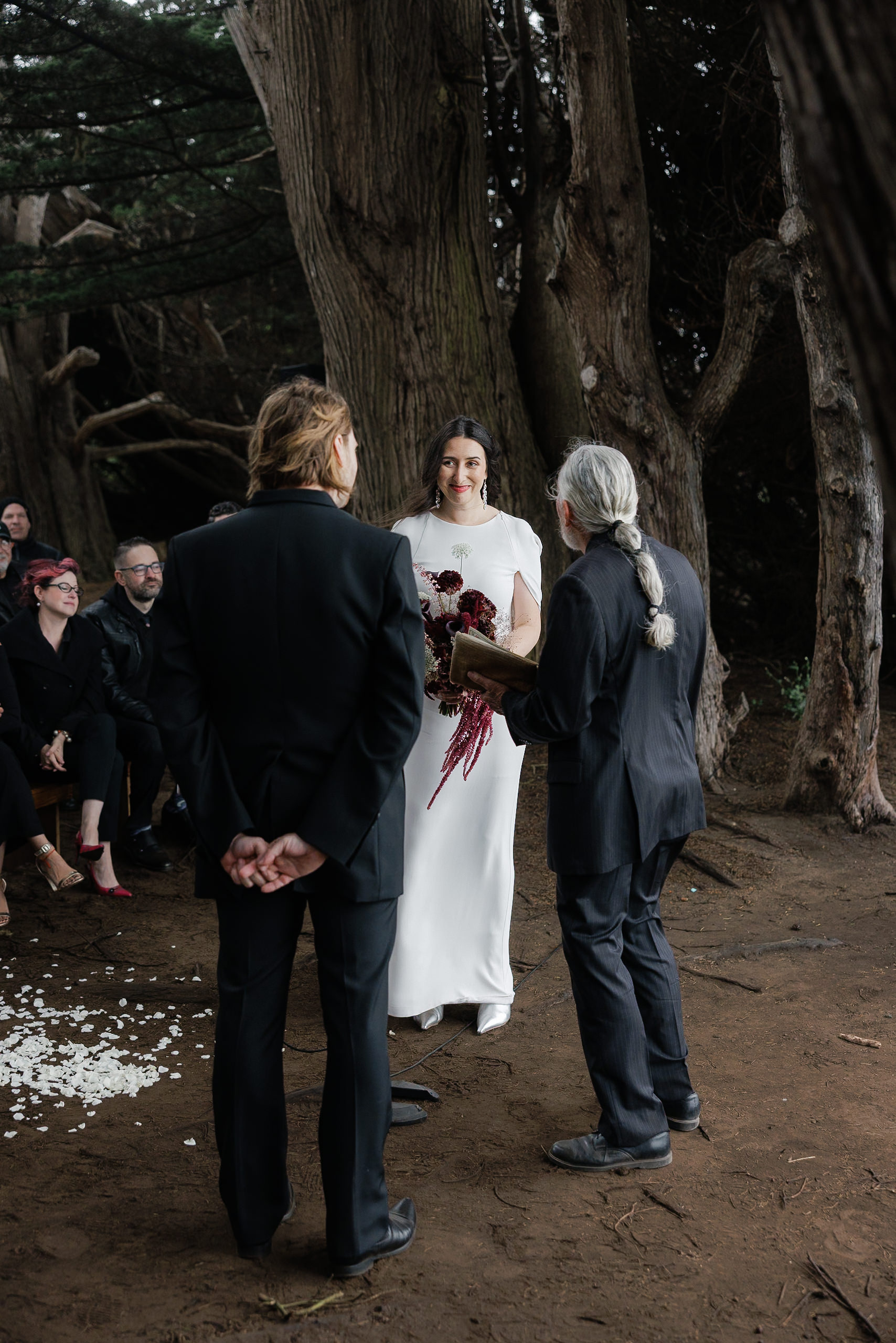 Bride at Mendocino forest altar with dark bouquet; officiant and guests stand amid scattered petals.