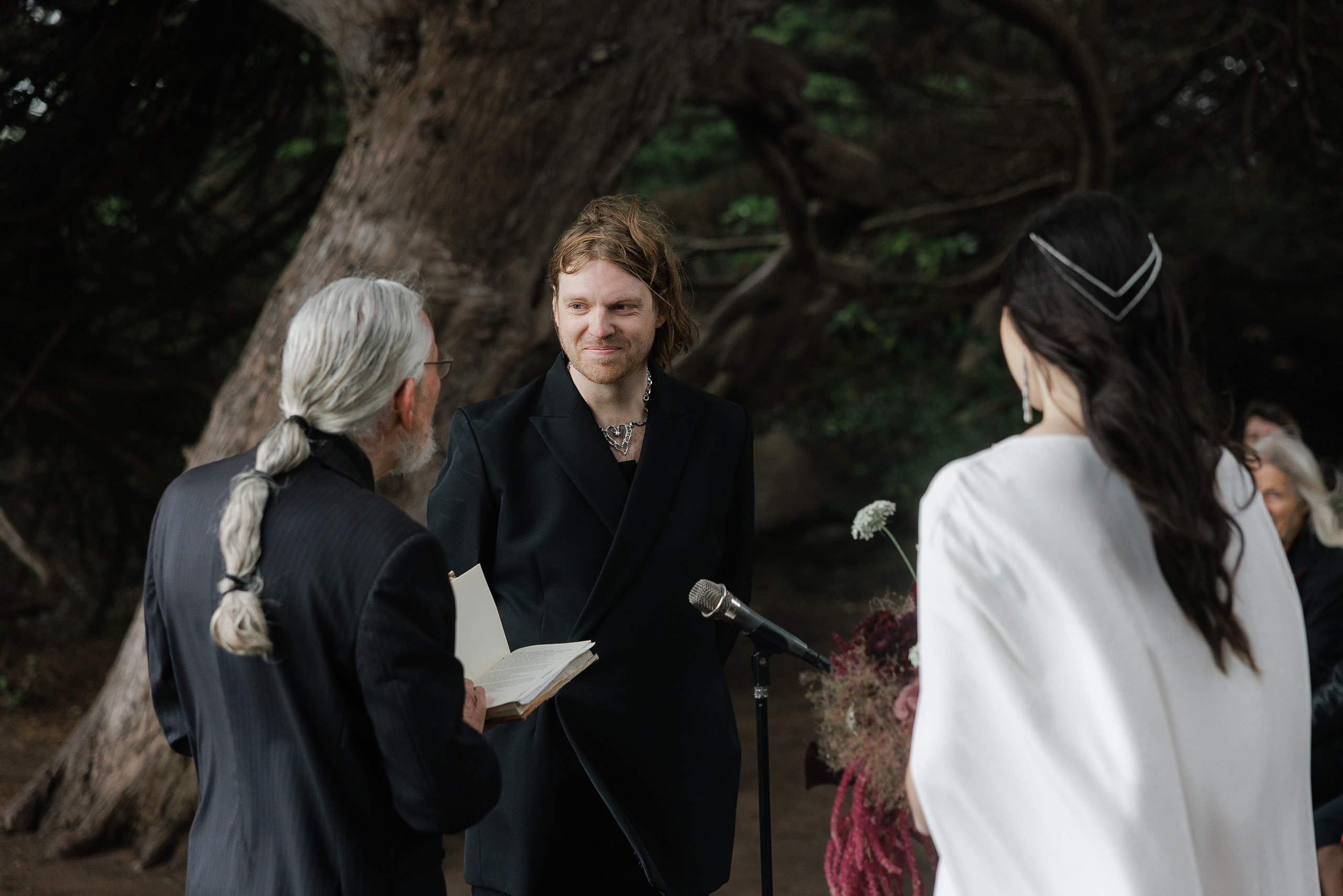 Mendocino Wedding: Outdoor Ceremony with Groom in Black Suit & Bride in White Dress Amidst Forest Backdrop.