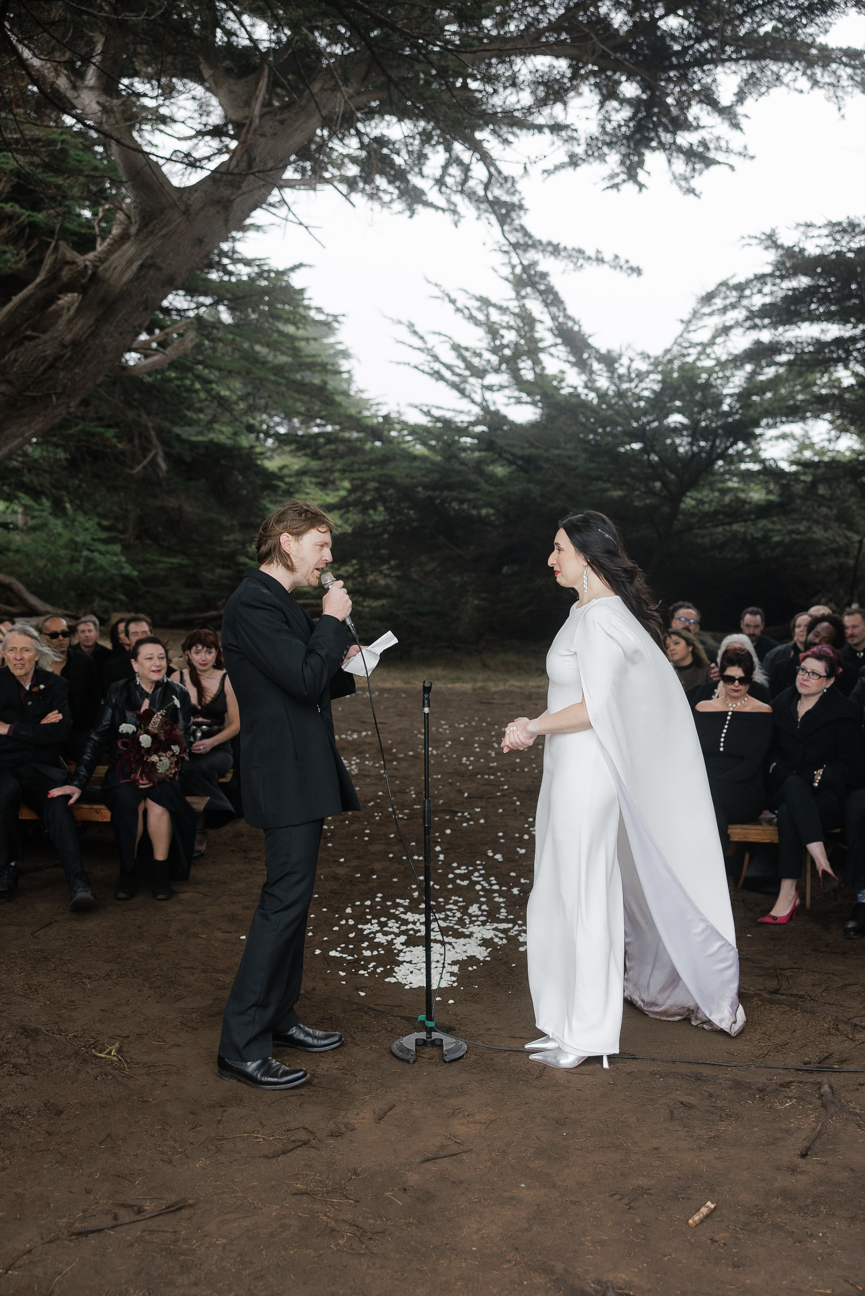 Outdoor Wedding in Mendocino: Bride in Cape & Groom Exchange Vows Beneath Tall Trees.