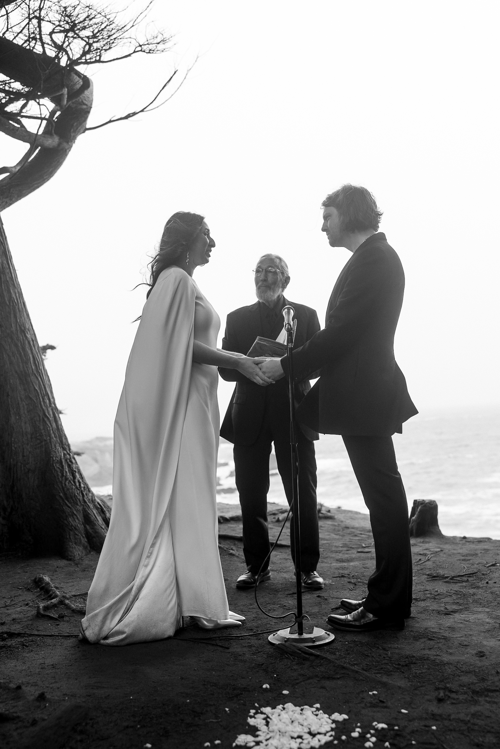 Black and white Mendocino beach wedding ceremony, bride & groom holding hands, surrounded by ocean views.