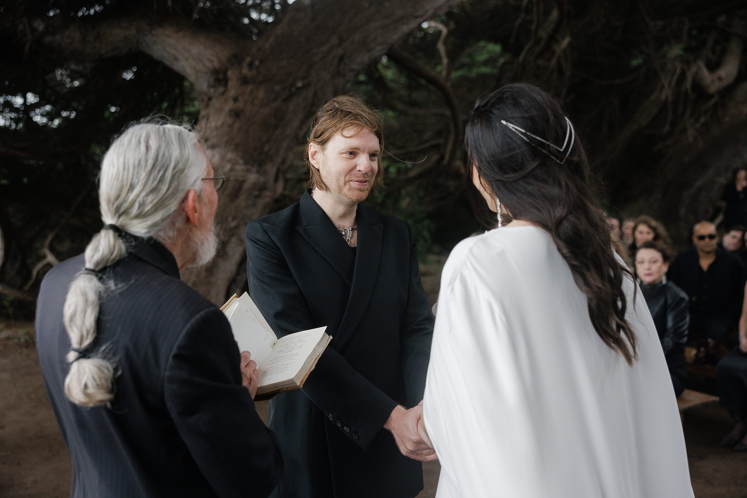 Elegant outdoor Mendocino wedding: bride in white, groom in dark suit, officiant under twisting trees.