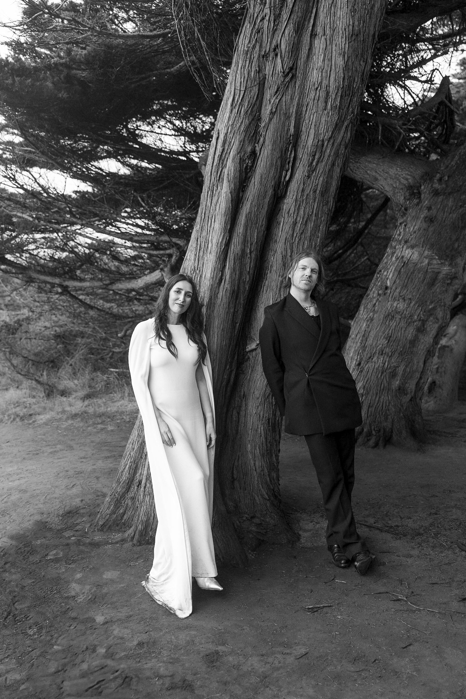 Elegant black-and-white wedding photo: couple under Mendocino trees, bride in white dress, groom in dark suit.