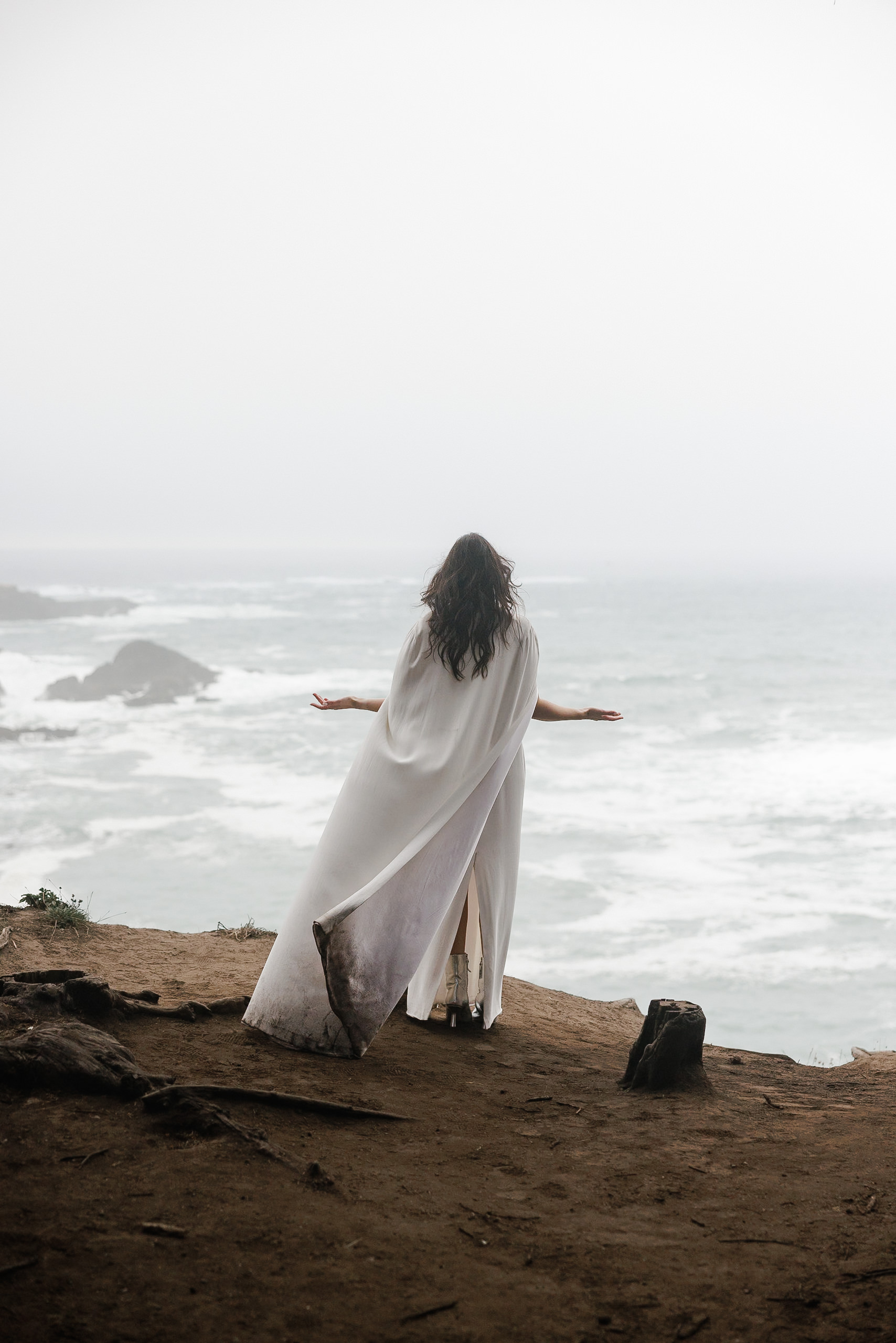 Elegant bride in flowing white cloak atop Mendocino cliffs, ocean backdrop.