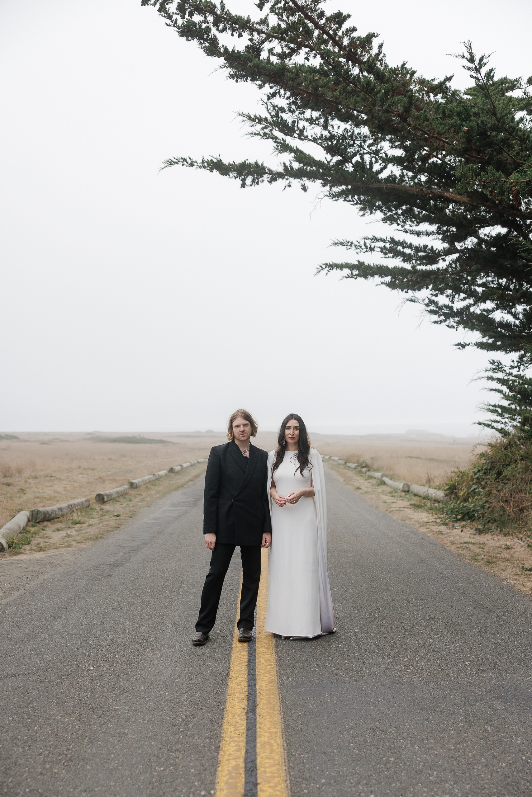 Elegant Wedding Photography in Mendocino: Couple on Scenic Road with Striking Tree Under Clouded Sky.