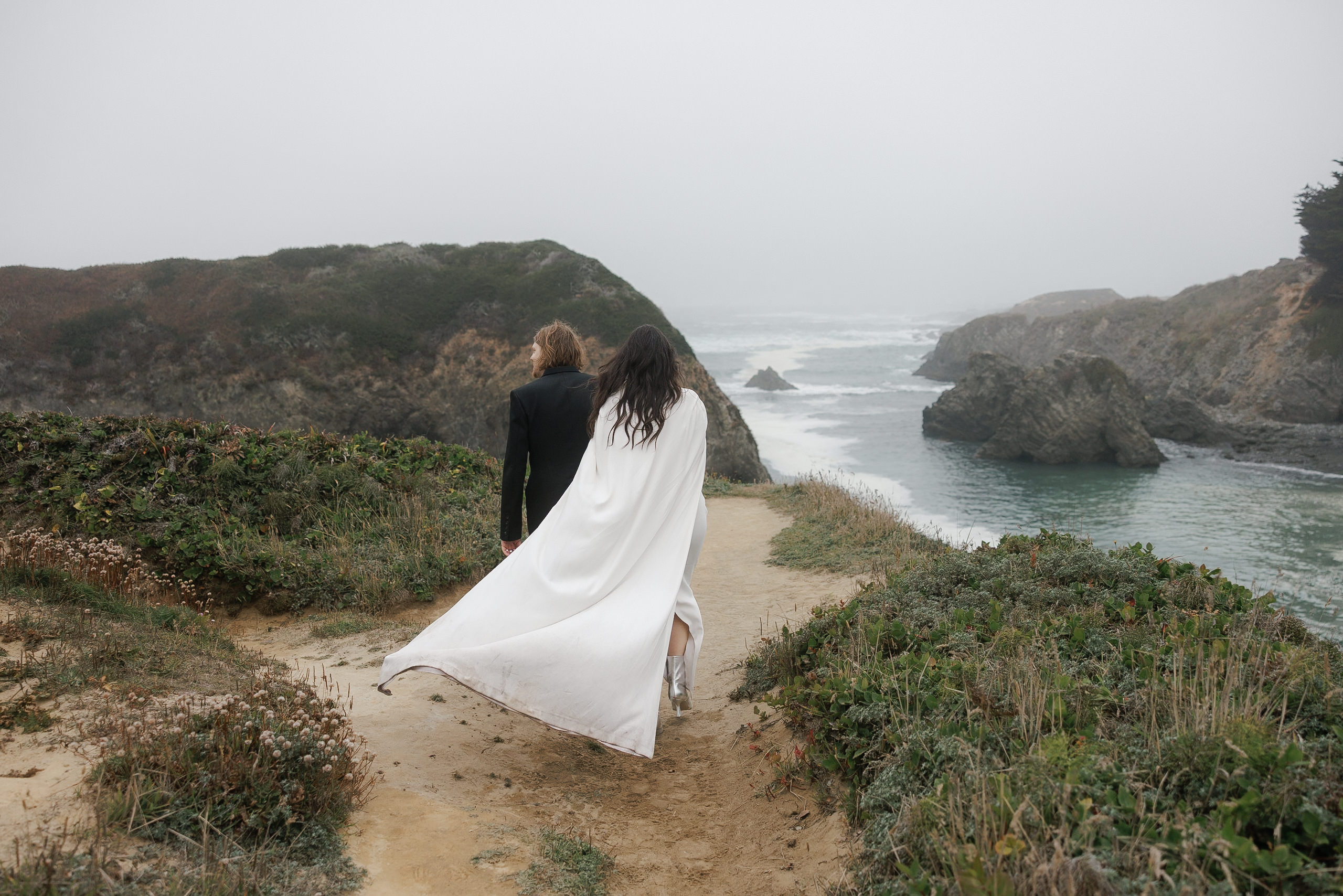 Elegant Wedding Stroll: Black Suit & White Cloak by Mendocino Cliffs, Moody Overcast Backdrop.