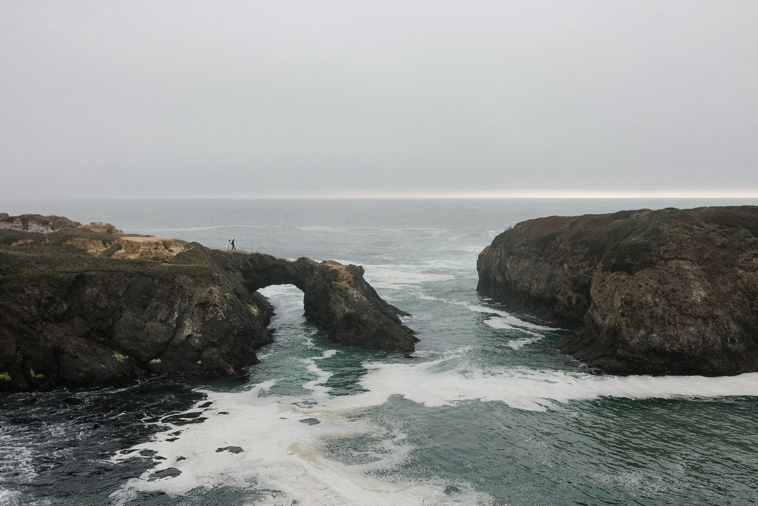 Elegant bride on Mendocino rock arch, enveloped by ocean waves and misty coastal cliffs.