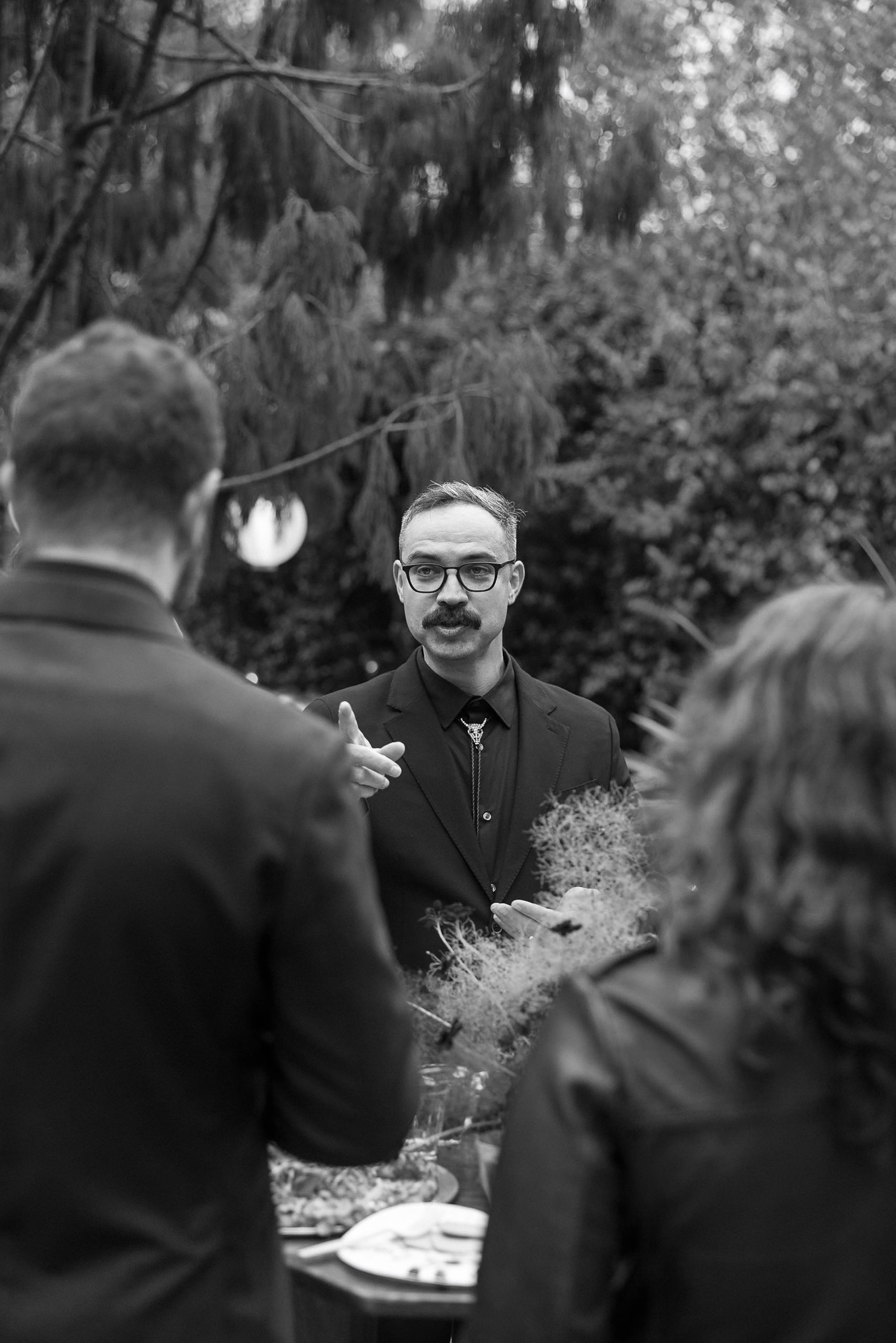 Black and white outdoor conversation in Mendocino; mustached man in glasses speaks to two others amidst lush greenery.