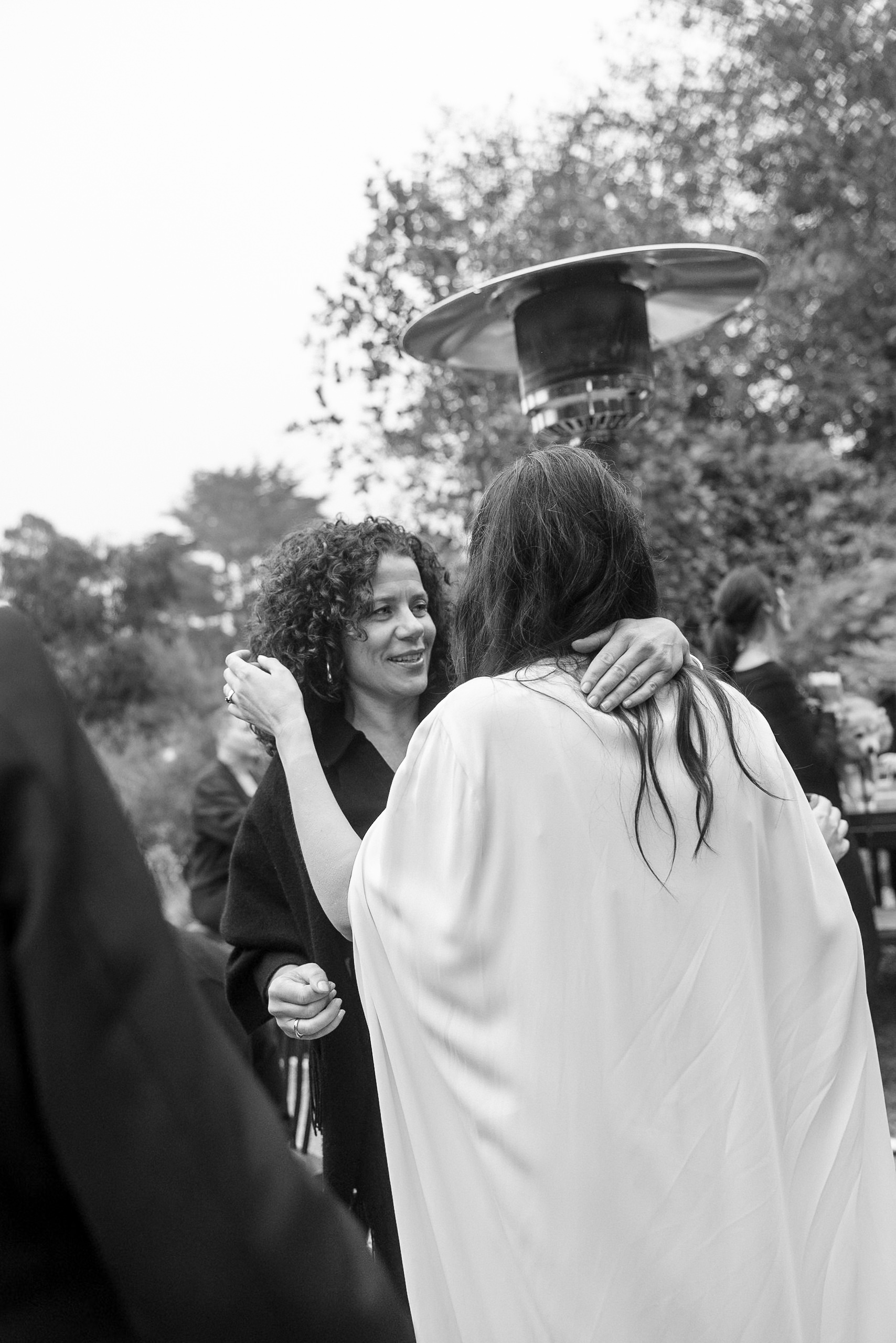 Black & White Wedding Moment: Embrace Under Patio Heater, Smiling with Mendocinos Greenery Background.