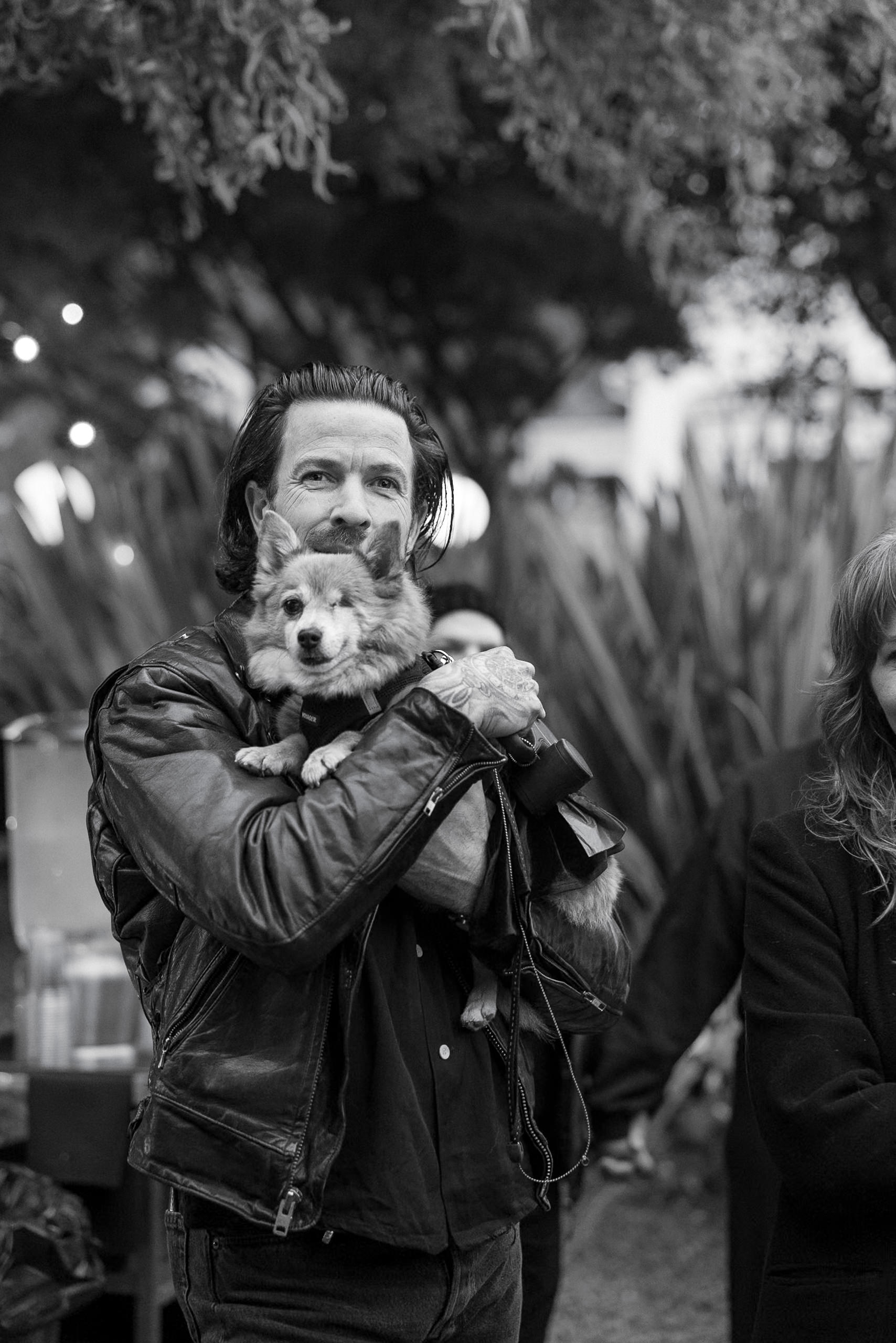 Black-and-white photo: person in leather jacket holding corgi mix amid Mendocinos rustic charm. Trees and string lights aglow.