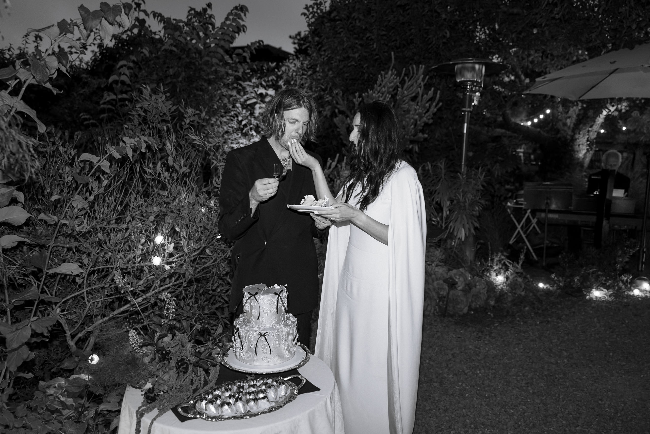 Black & White Elegant Wedding in Mendocino Garden: Cake Sharing Moment Under String Lights.