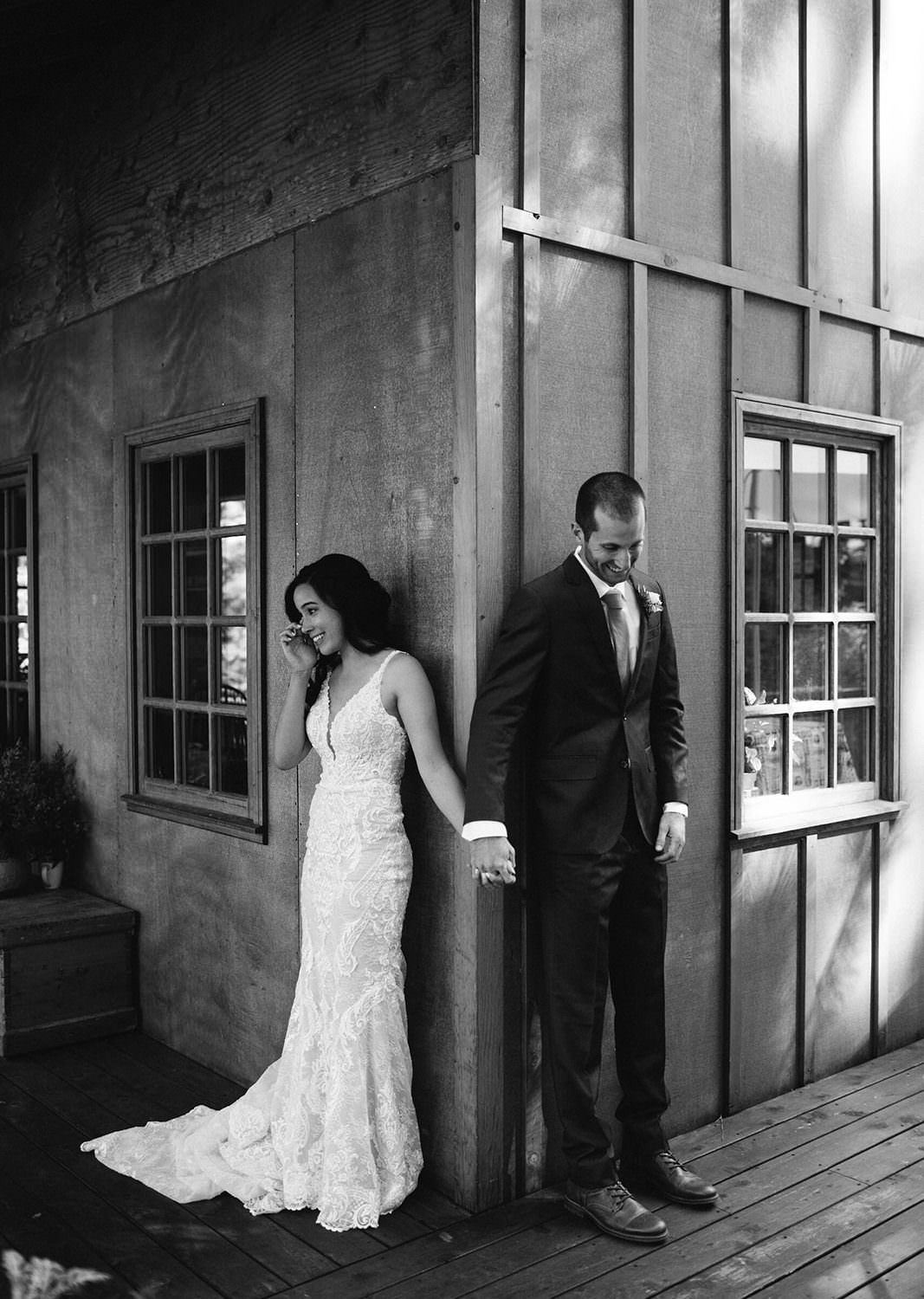 Bride in lace gown, groom in suit hold hands, outdoor corner setting with wooden walls and soft shadows.