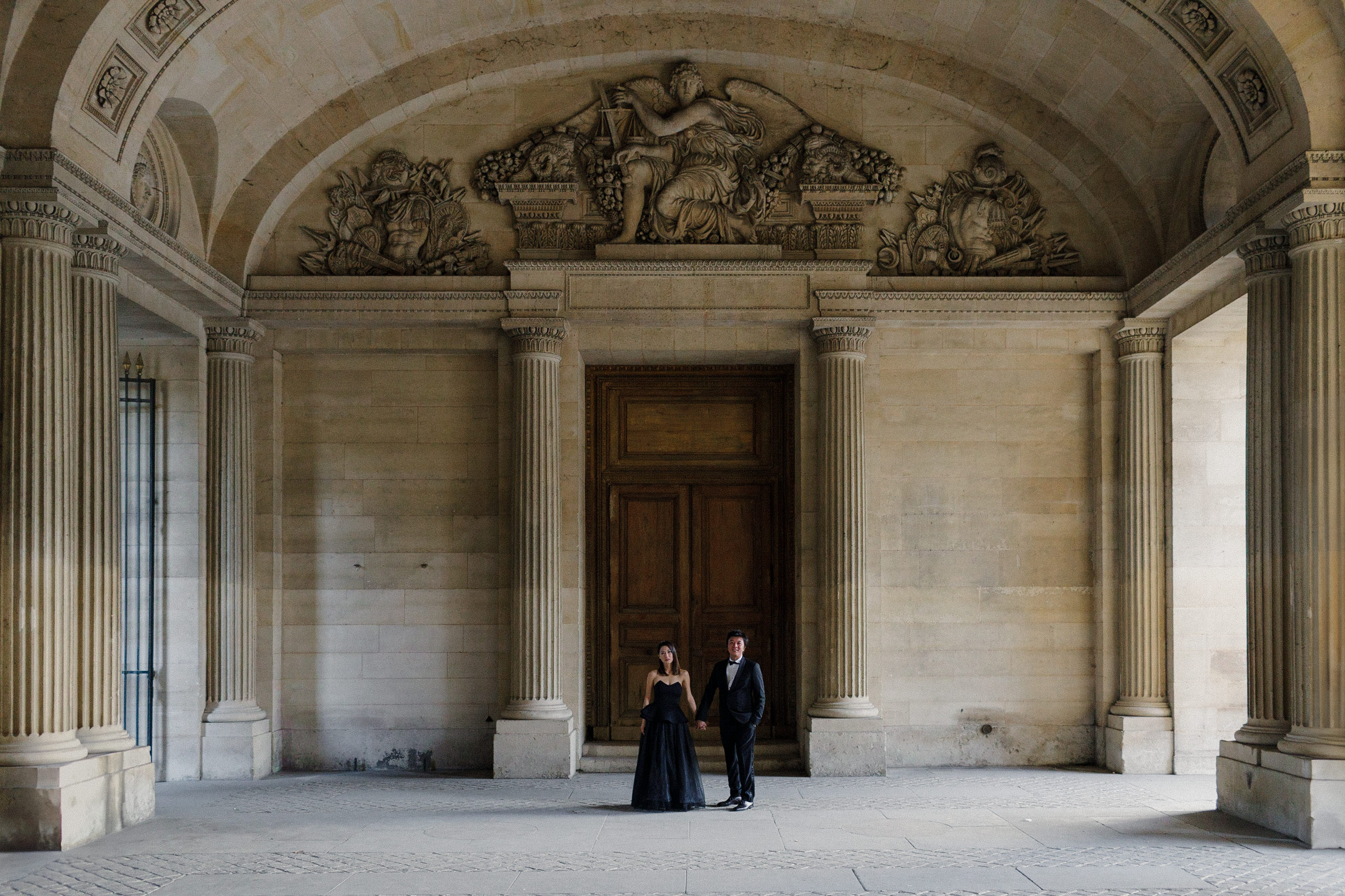 Elegant couple in a grand hallway with ornate carvings and columns; black dress and suit.