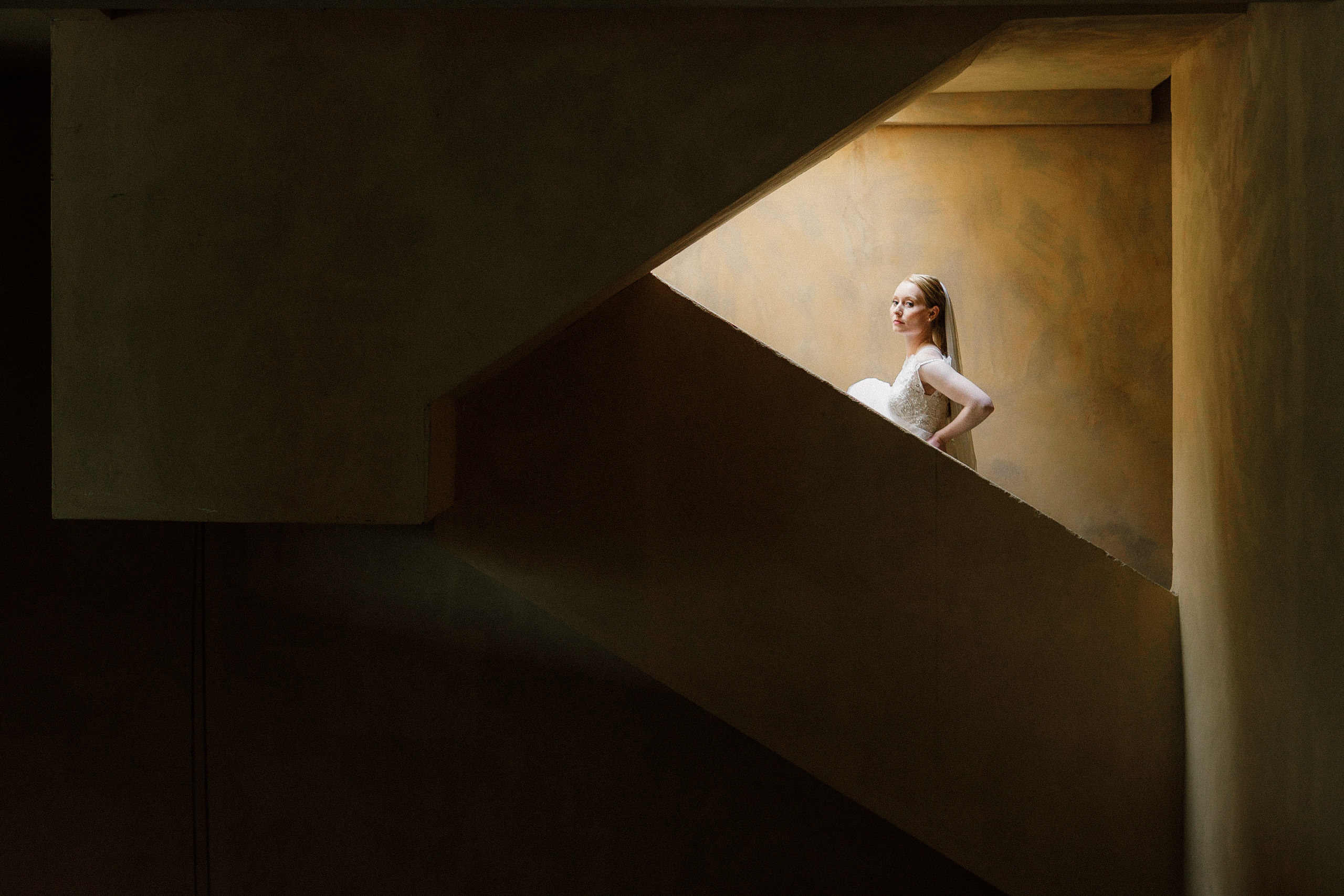 Elegant bride in white dress amid sunlit geometric stairwell, showcasing modern wedding photography artistry.