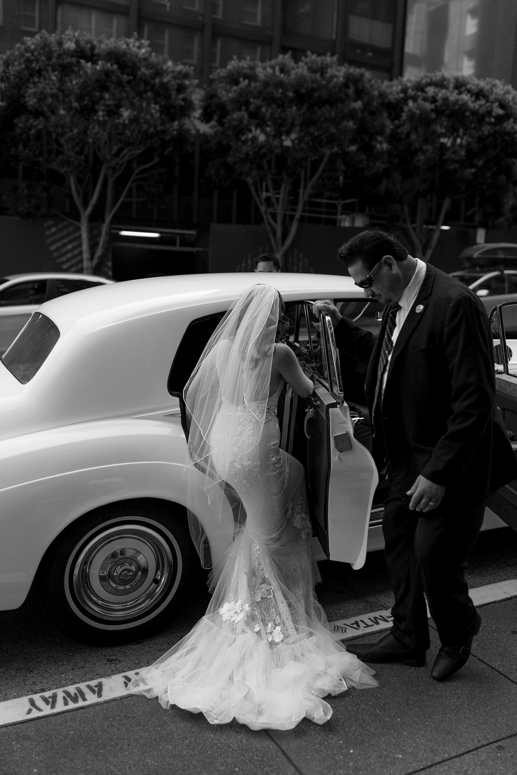 Elegant urban wedding: Bride in flowing gown steps into classic car; black and white photography.