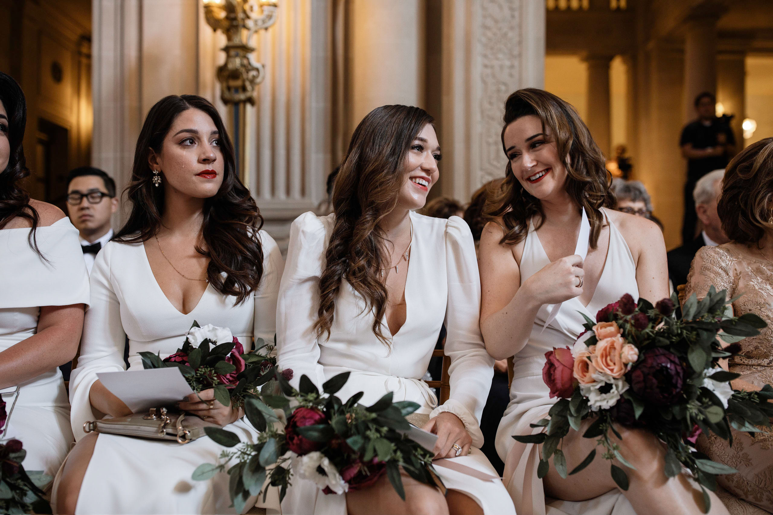 Elegant bridesmaids in white, holding floral bouquets at a joyful wedding reception.