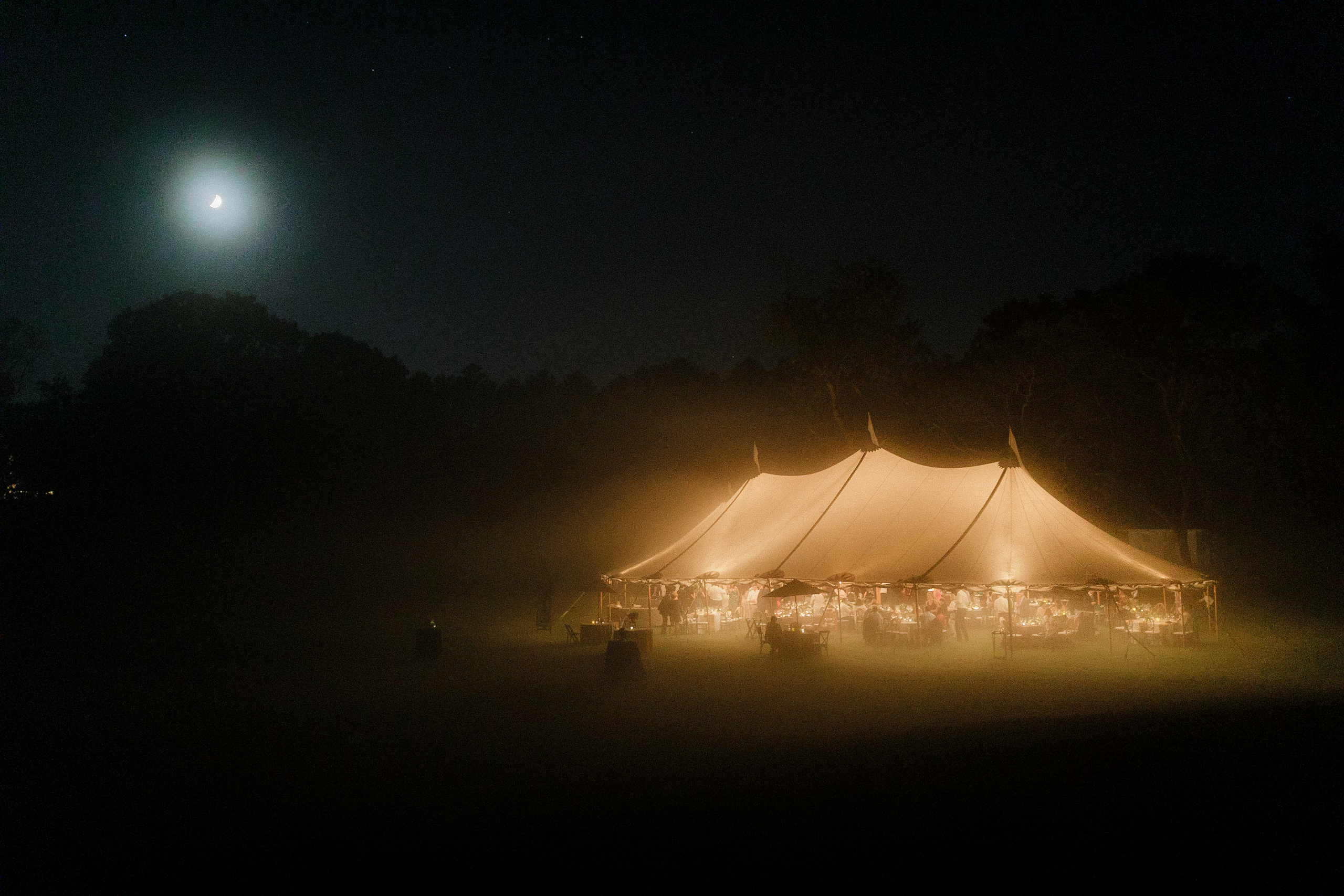 Crescent moon wedding tent lit warmly, serene night, glowing event, silhouetted trees backdrop.
