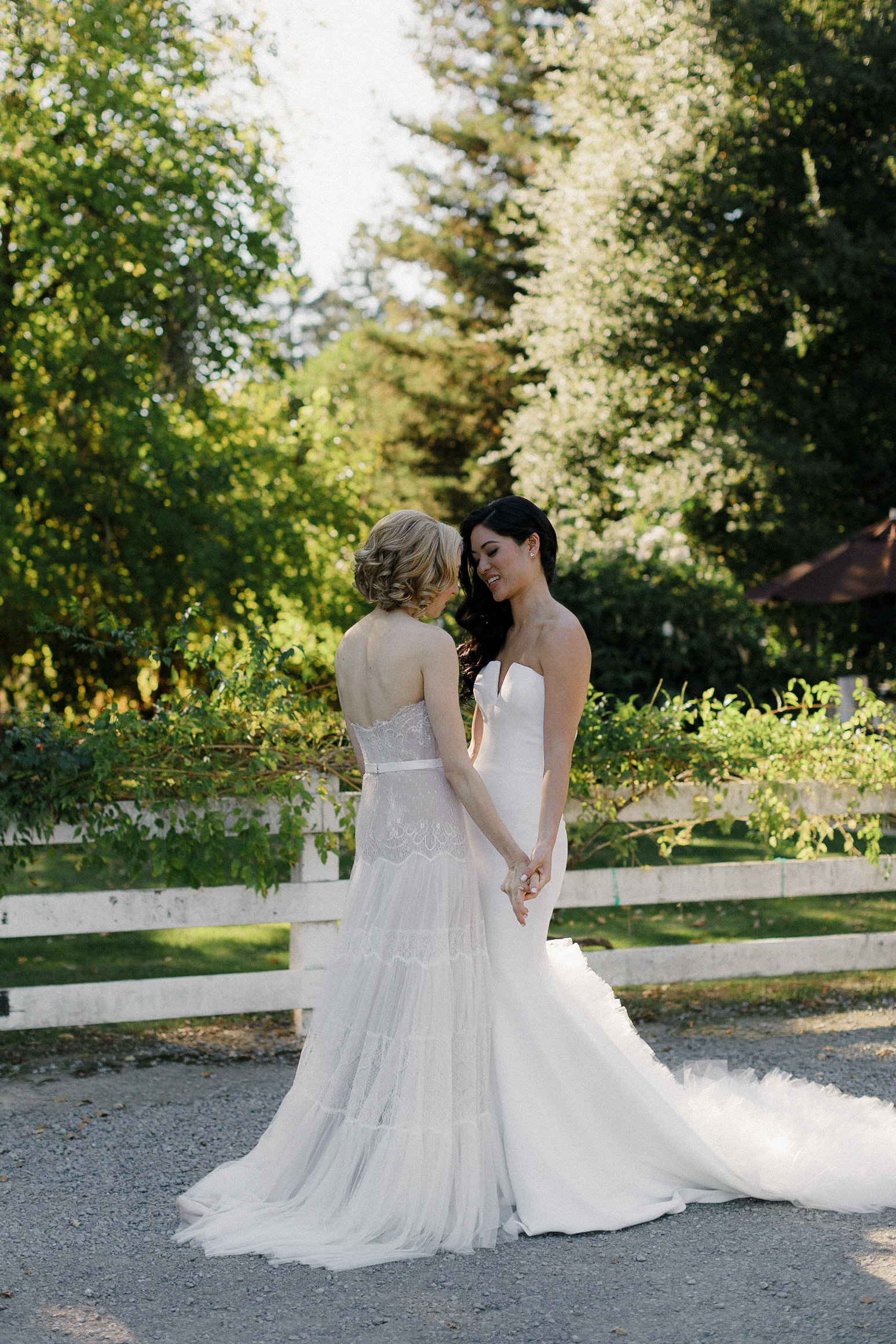 Elegant Brides in White Gowns Holding Hands by Rustic Fence | Serene Outdoor Wedding Photography.