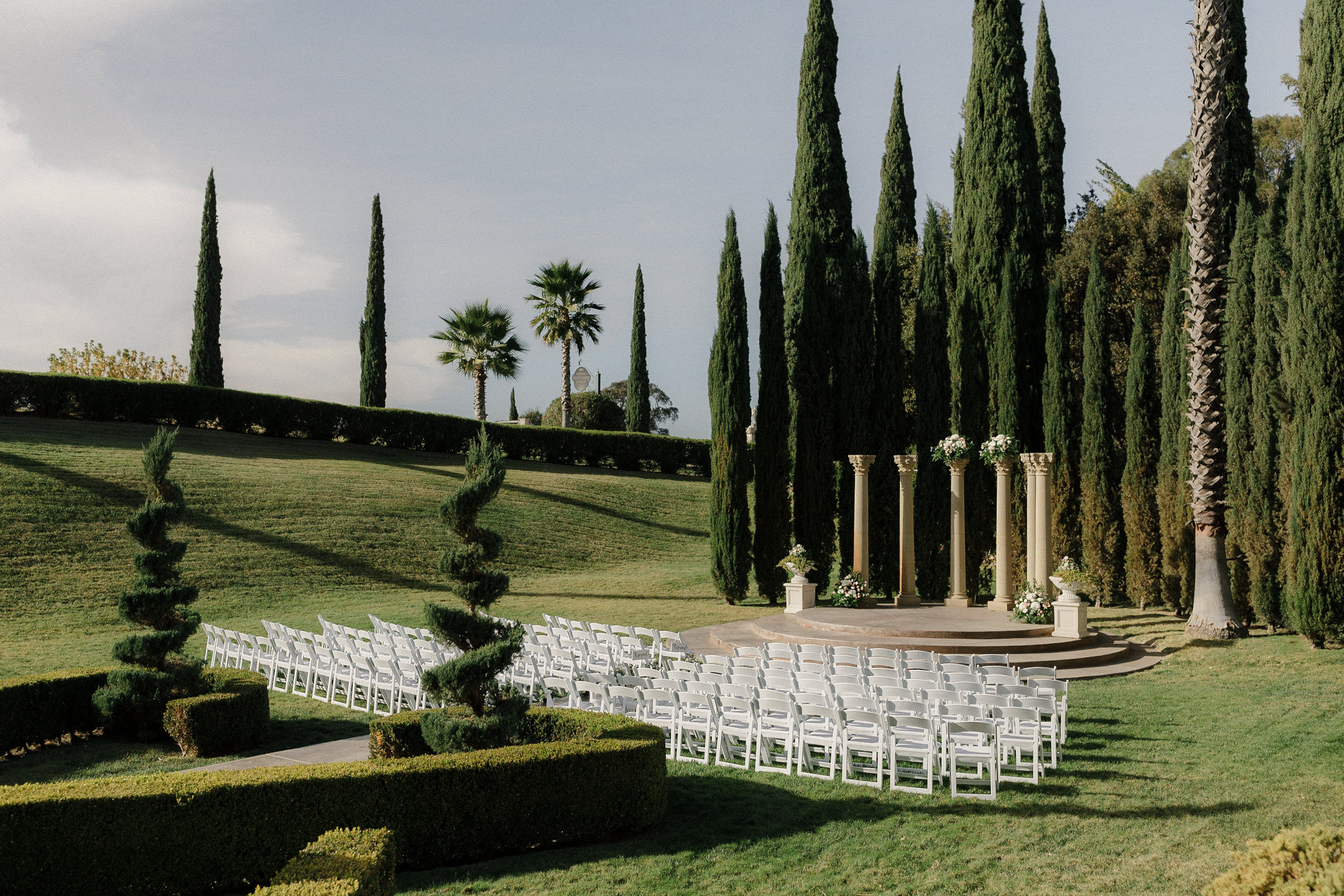 Elegant Outdoor Garden Wedding with White Chairs & Floral Columns Amidst Cypress & Palm Trees.