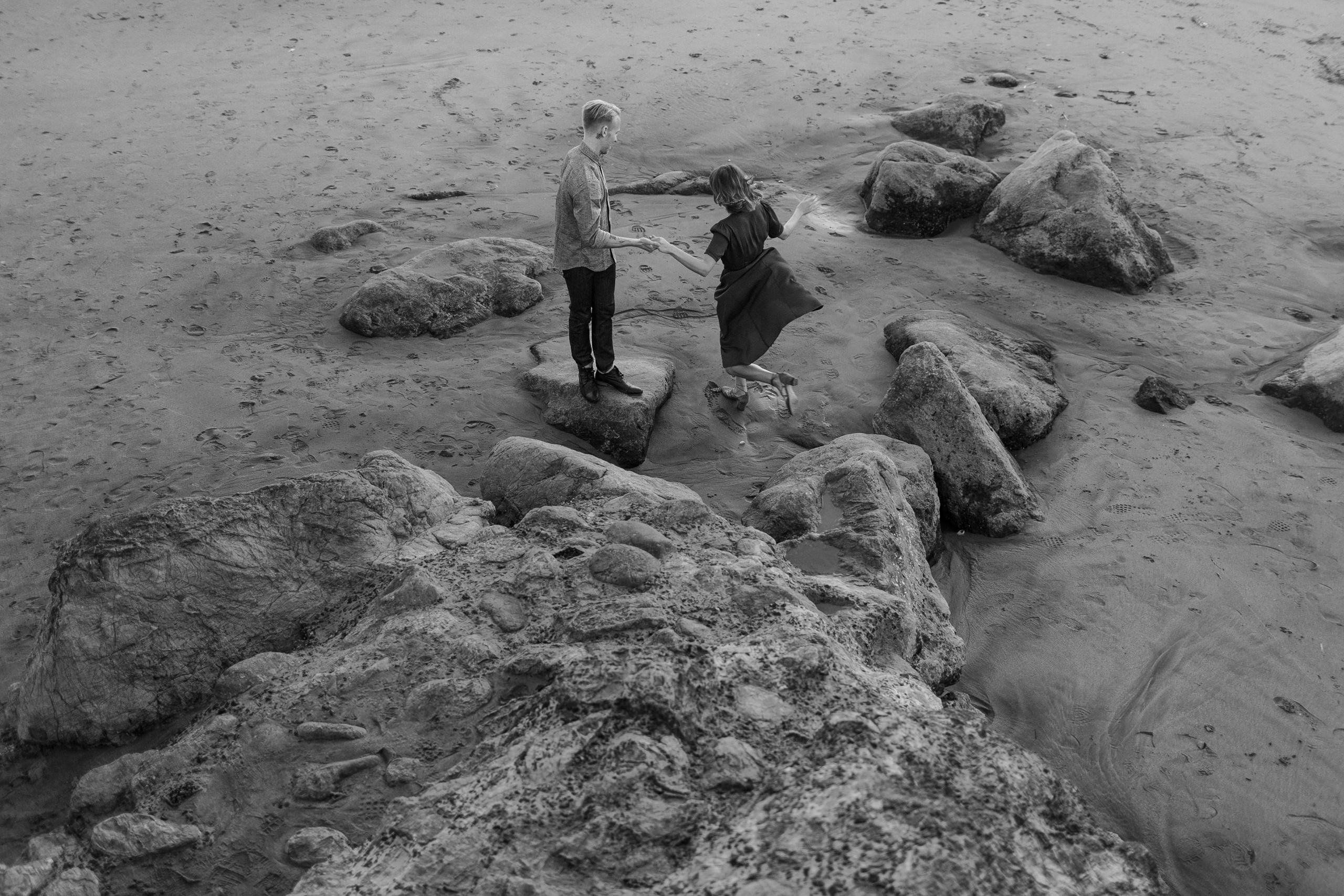 Black & white engagement photo: couple holding hands on beach rocks, timeless elegance.