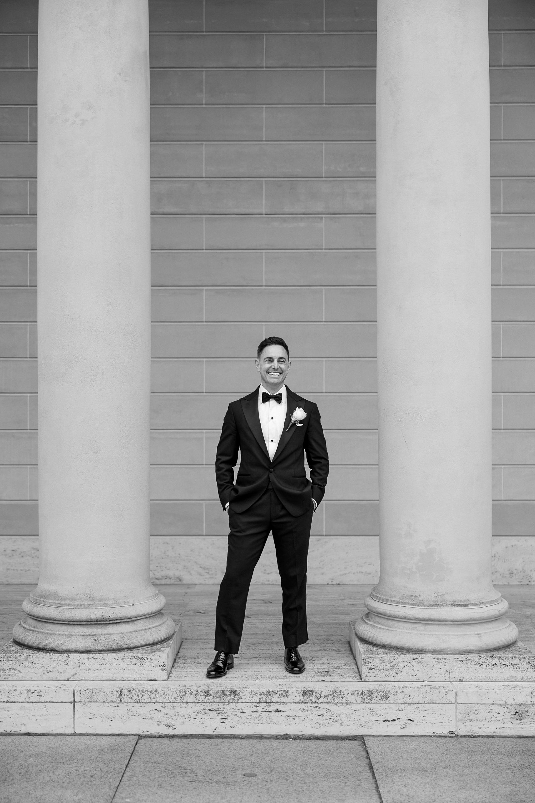 Elegant groom in tuxedo framed by stone columns on a terrace; black and white portrait.