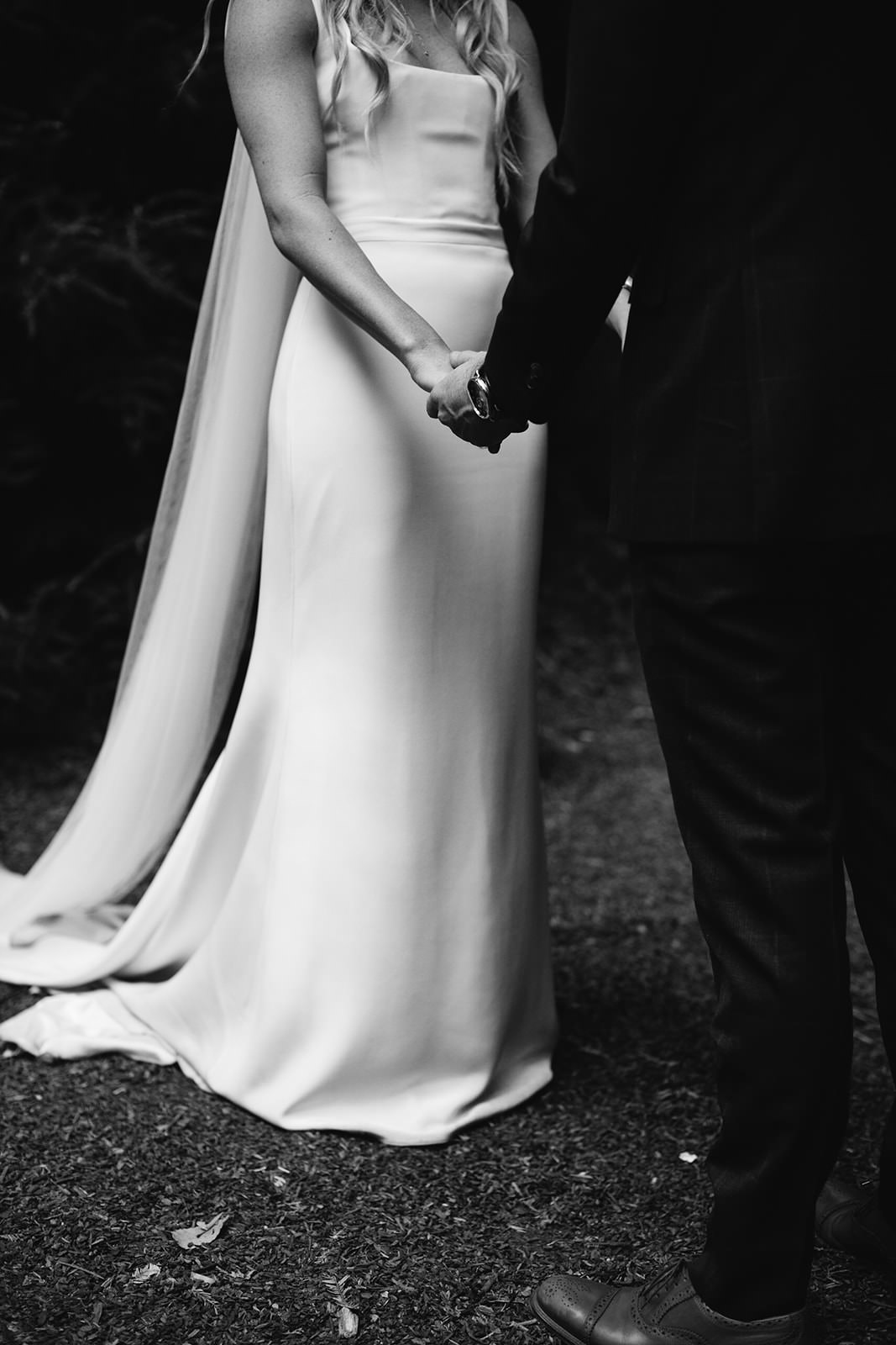 A bride and groom hold hands, exuding timeless elegance amidst lush California foliage in a romantic black-and-white scene.