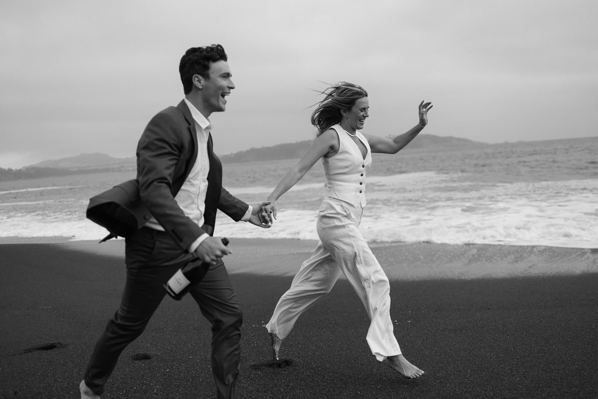 Elegant black and white beach wedding photo: couple joyfully runs barefoot, man in suit with wine, woman in white jumpsuit.