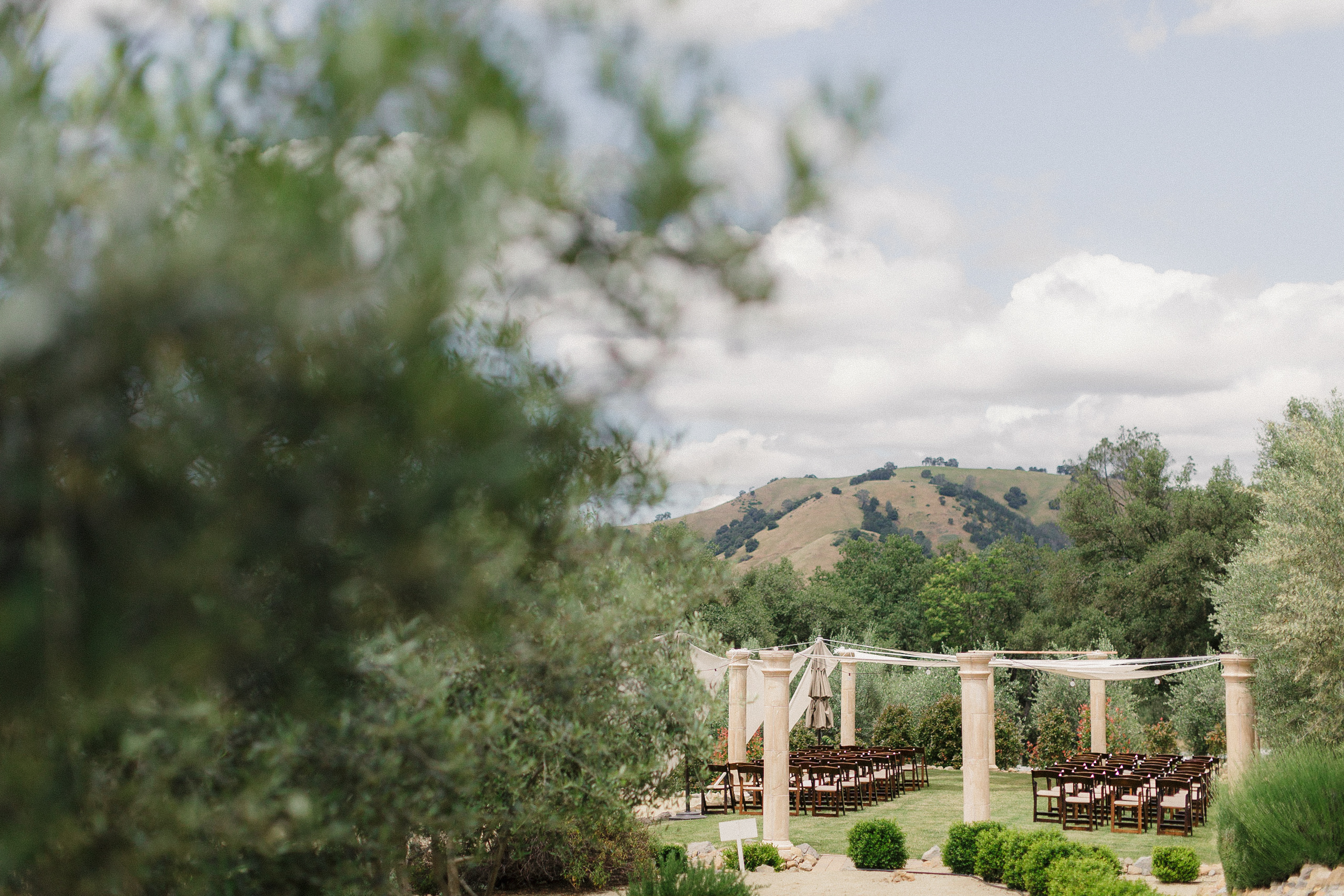 Elegant outdoor wedding ceremony; wooden chairs & trellis amidst hills, clouds, and lush greenery.
