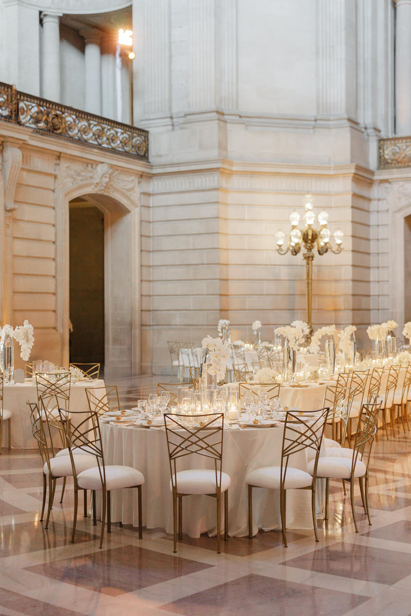 Lavish reception hall: white tablecloths, gold chairs, tall floral centers & ambient candlelight.