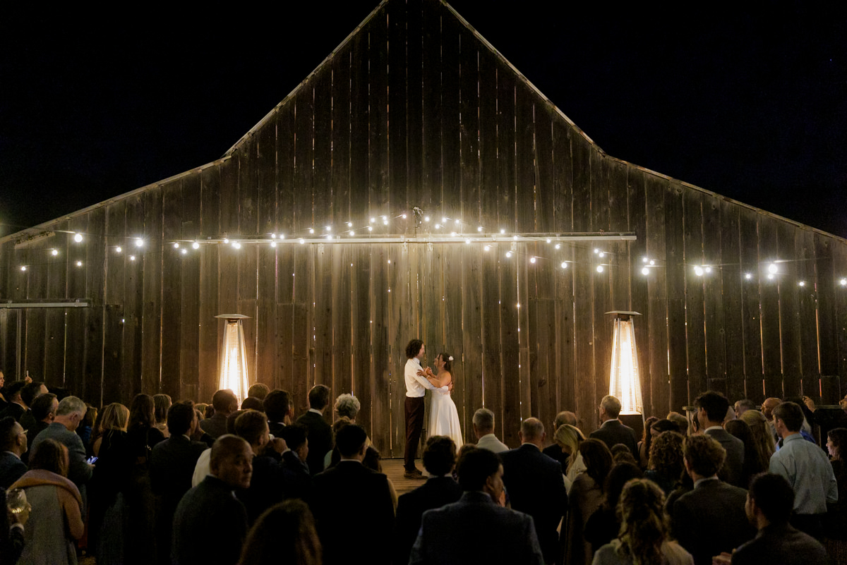 Enchanting Barn Wedding: Couples First Dance Under String Lights Amid Cherished Guests.