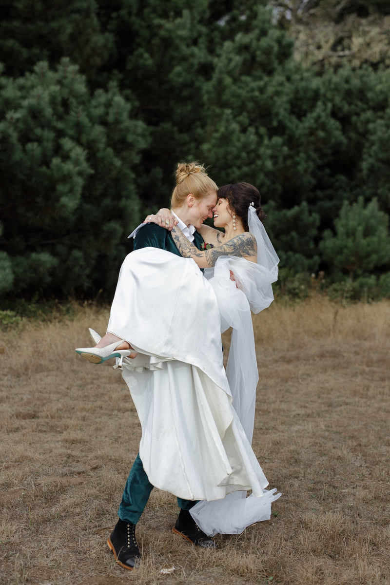 Elegant bride in a white dress lovingly embraced by groom in a green suit amidst lush greenery.
