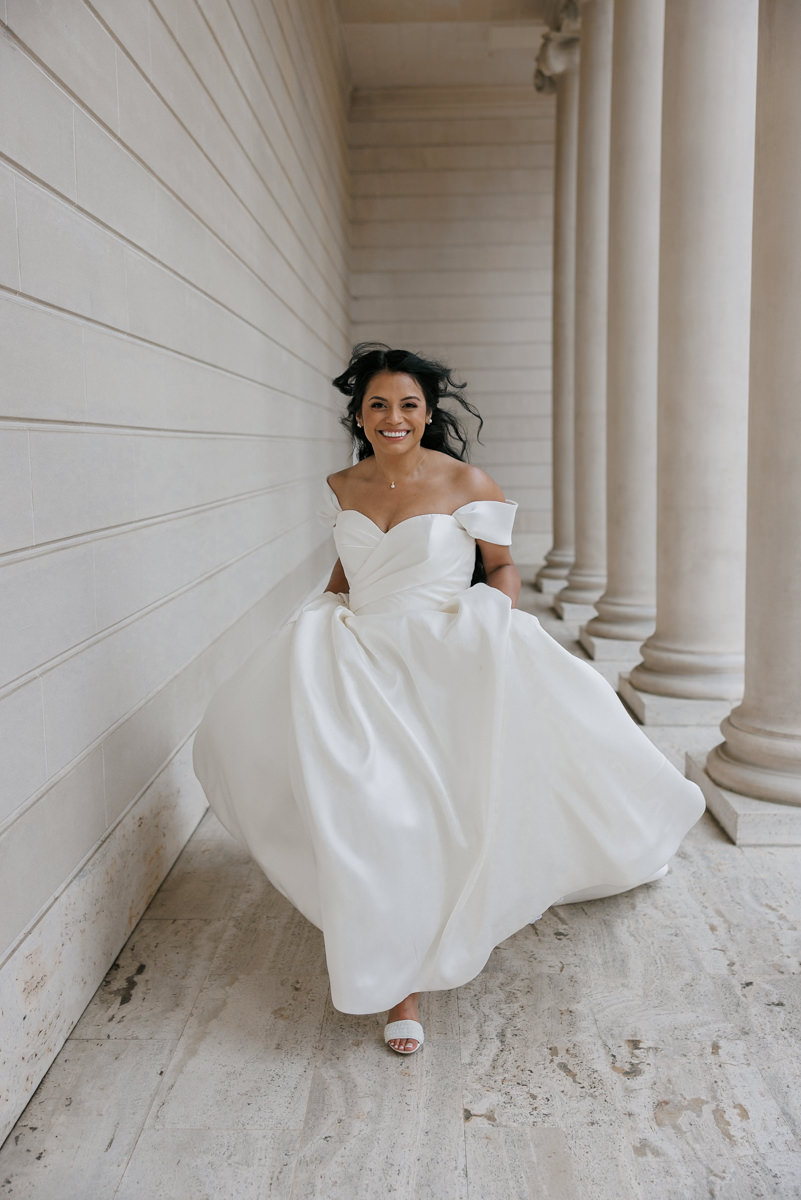 Elegant bride in flowing off-shoulder white dress, stone walkway, tall columns background.