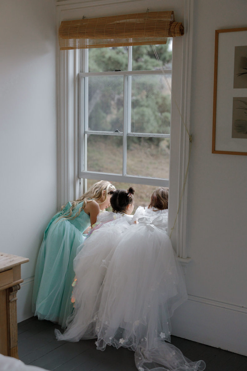 Pastel-clad bridesmaids daydream by window; soft greenery view. Elegant wedding photography.
