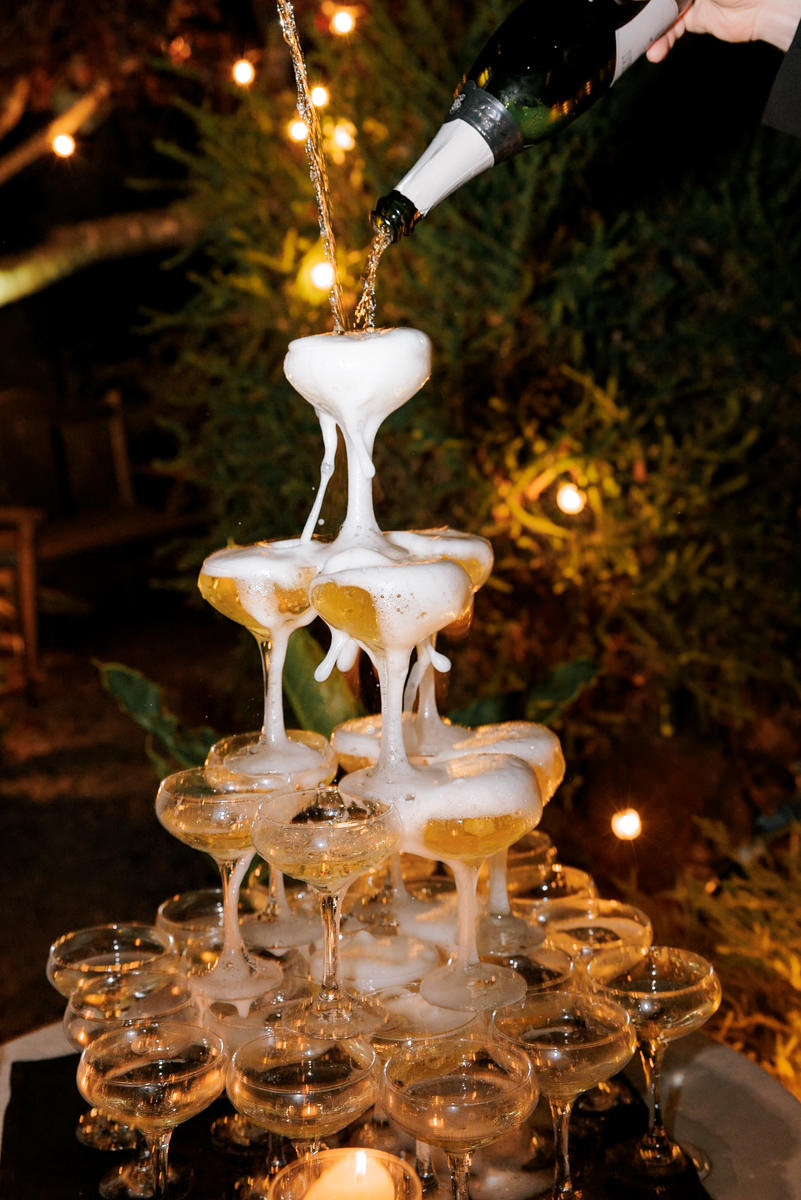 Champagne tower pour under glowing lights and greenery—luxury wedding photography.