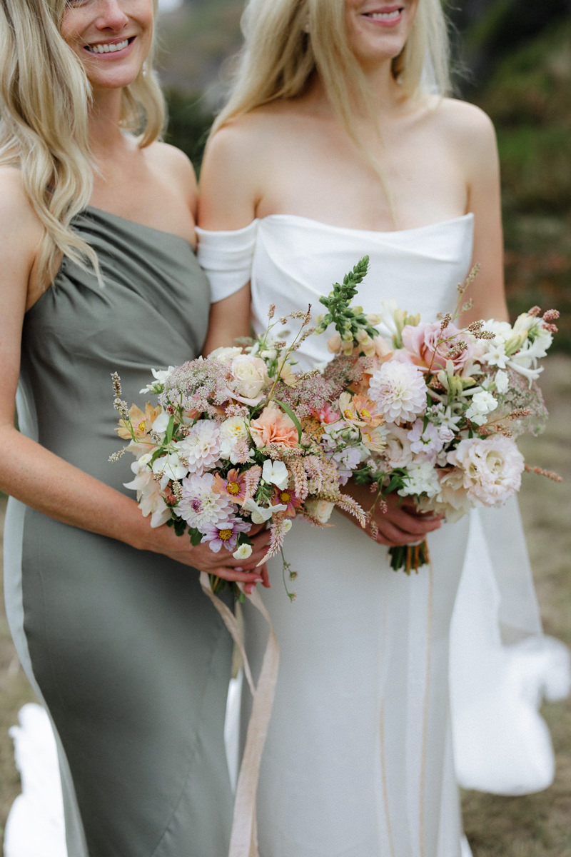 Elegant outdoor bridal portrait: bride in white off-shoulder dress, bridesmaid in sage-green gown, pink bouquets.