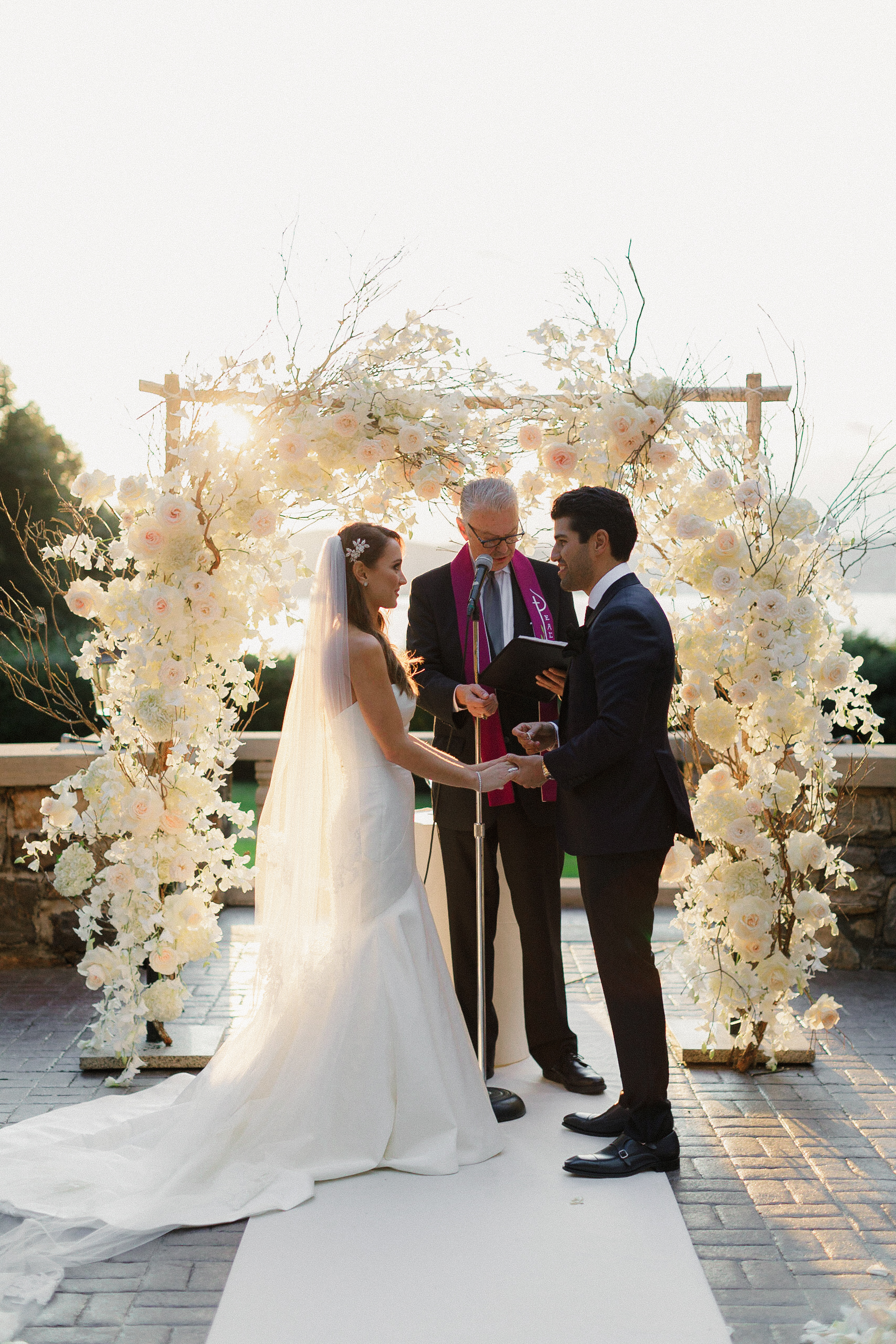 Elegant NYC Wedding Ceremony: Bride & Groom Exchange Vows Under Floral Arch in Sunlight.
