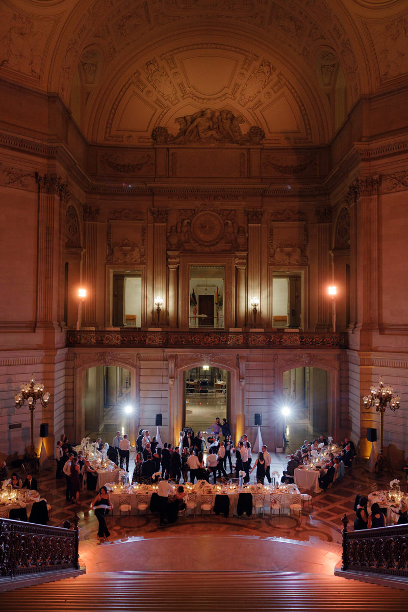 Elegantly dressed guests in a grand venue with ornate architecture, white linens, and soft lighting.