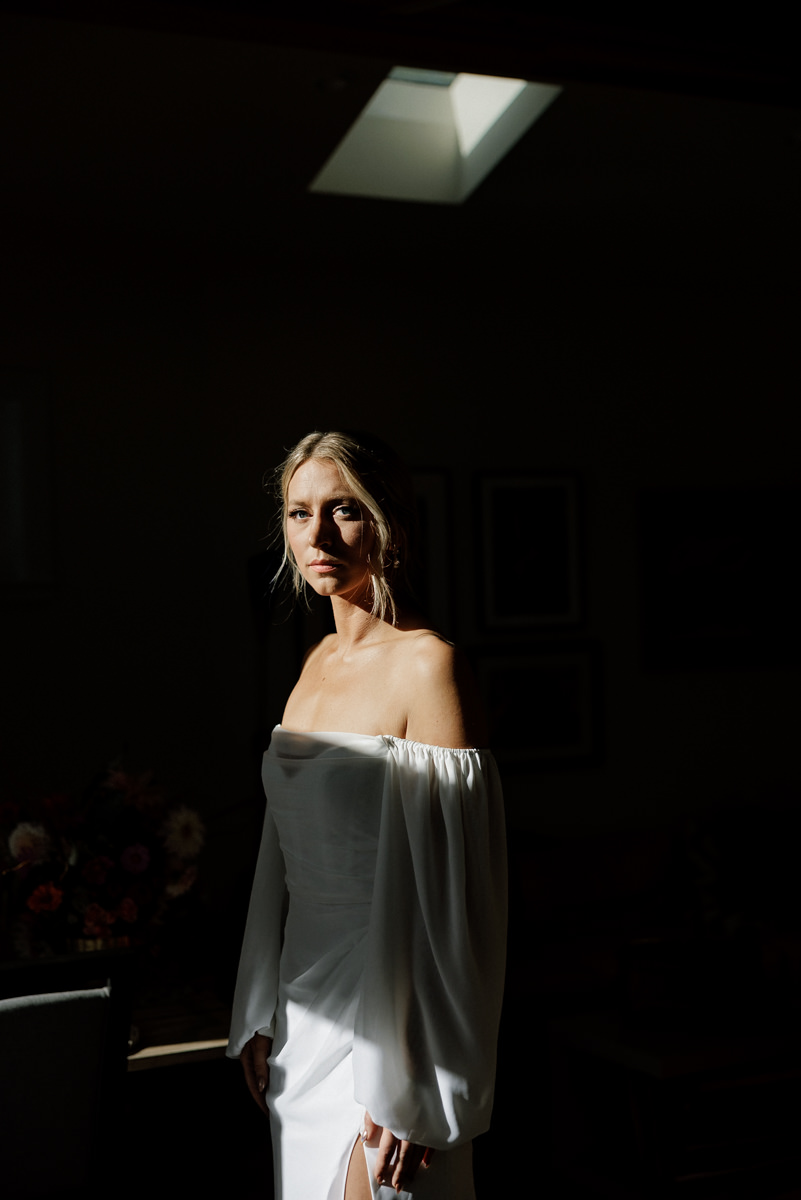 Elegant bride in off-the-shoulder dress, sunlight enhancing her poise, with bouquet backdrop.