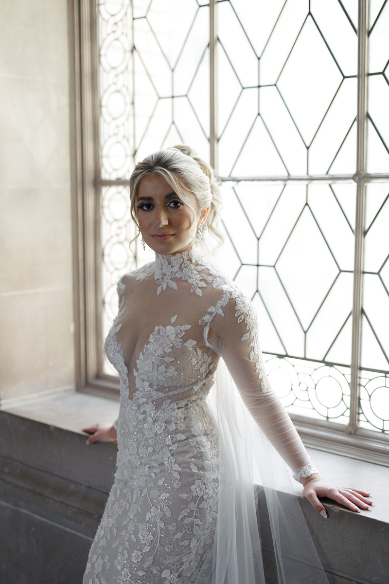 Bride in elegant lace wedding dress, near ornate window; soft natural light highlights her beauty.