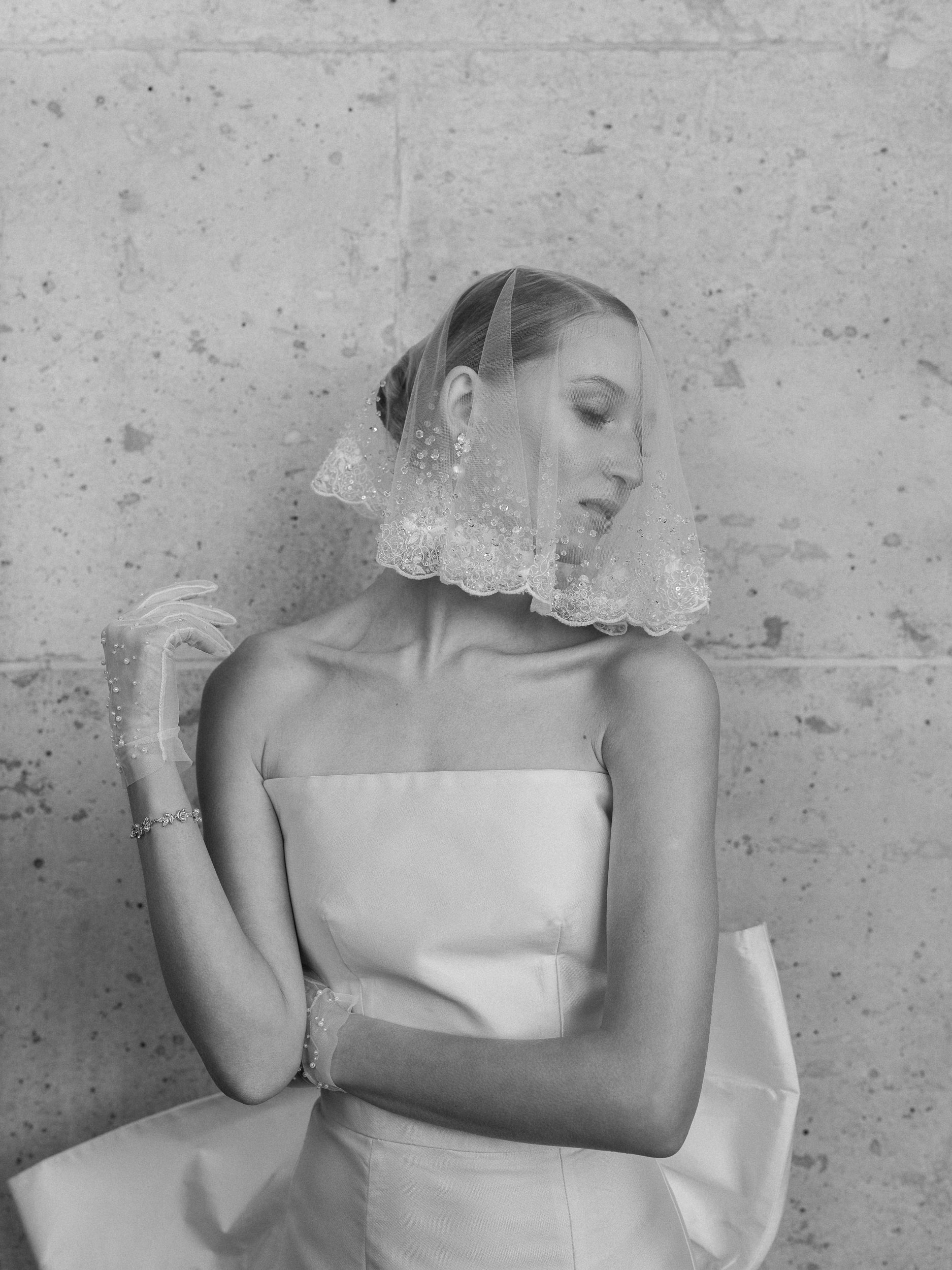 Woman in strapless wedding gown poses by textured wall, wearing pearl-detailed veil and sheer gloves, looking serenely to the side.