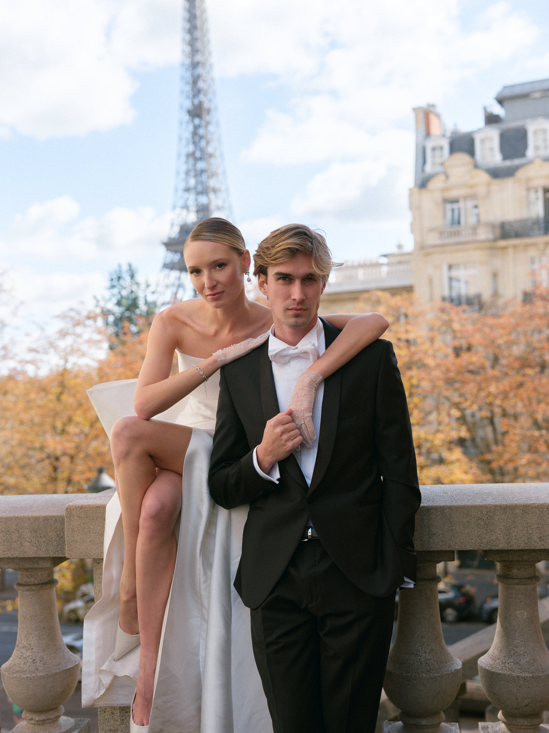 Bride in strapless white gown sits beside groom in black tuxedo; Eiffel Tower and Paris buildings visible on an autumn day.