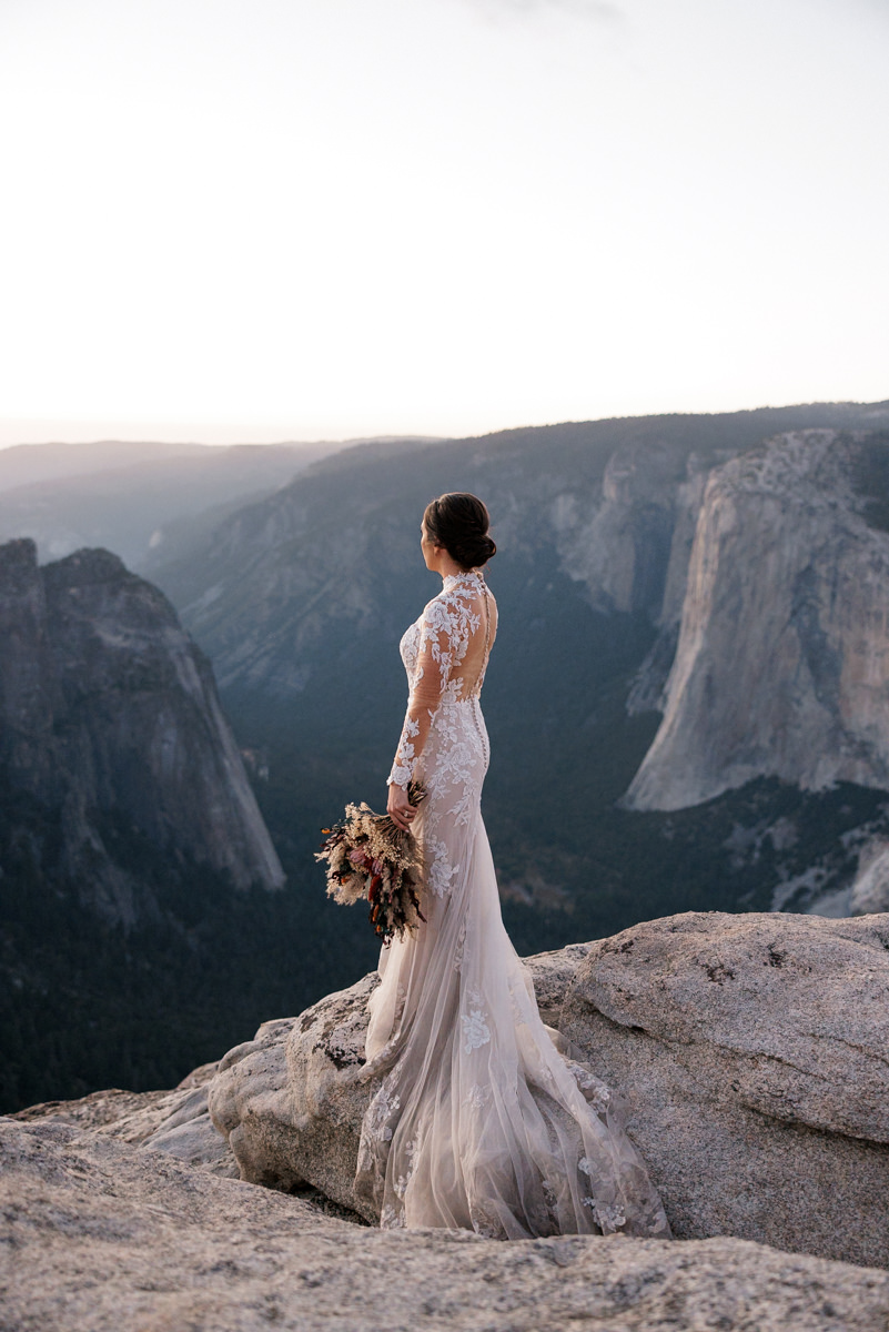 Elegant bride in lace gown gazes over mountain valley at sunset, bouquet in hand, glowing landscape.