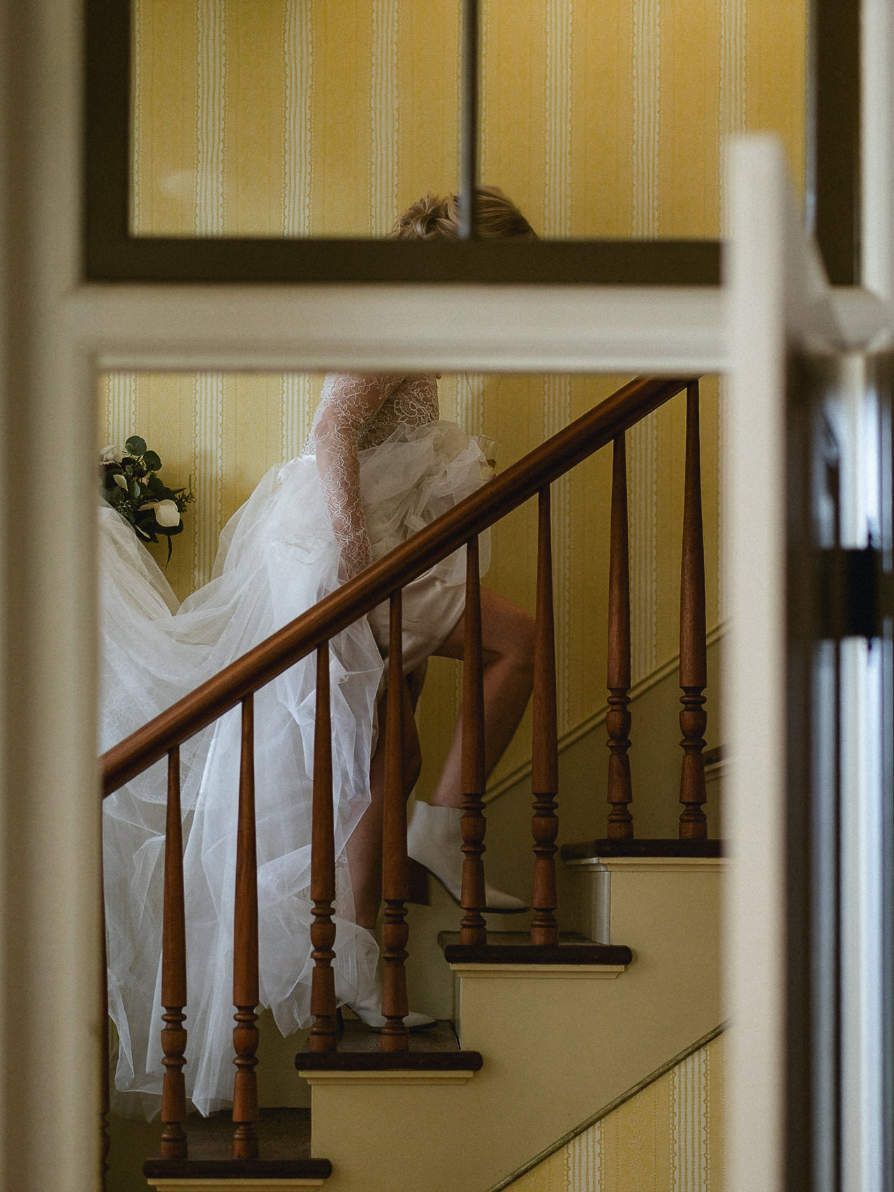Person in white wedding dress with bouquet walks up wooden stairs, yellow patterned wallpaper, seen from behind in Mendocino, CA.