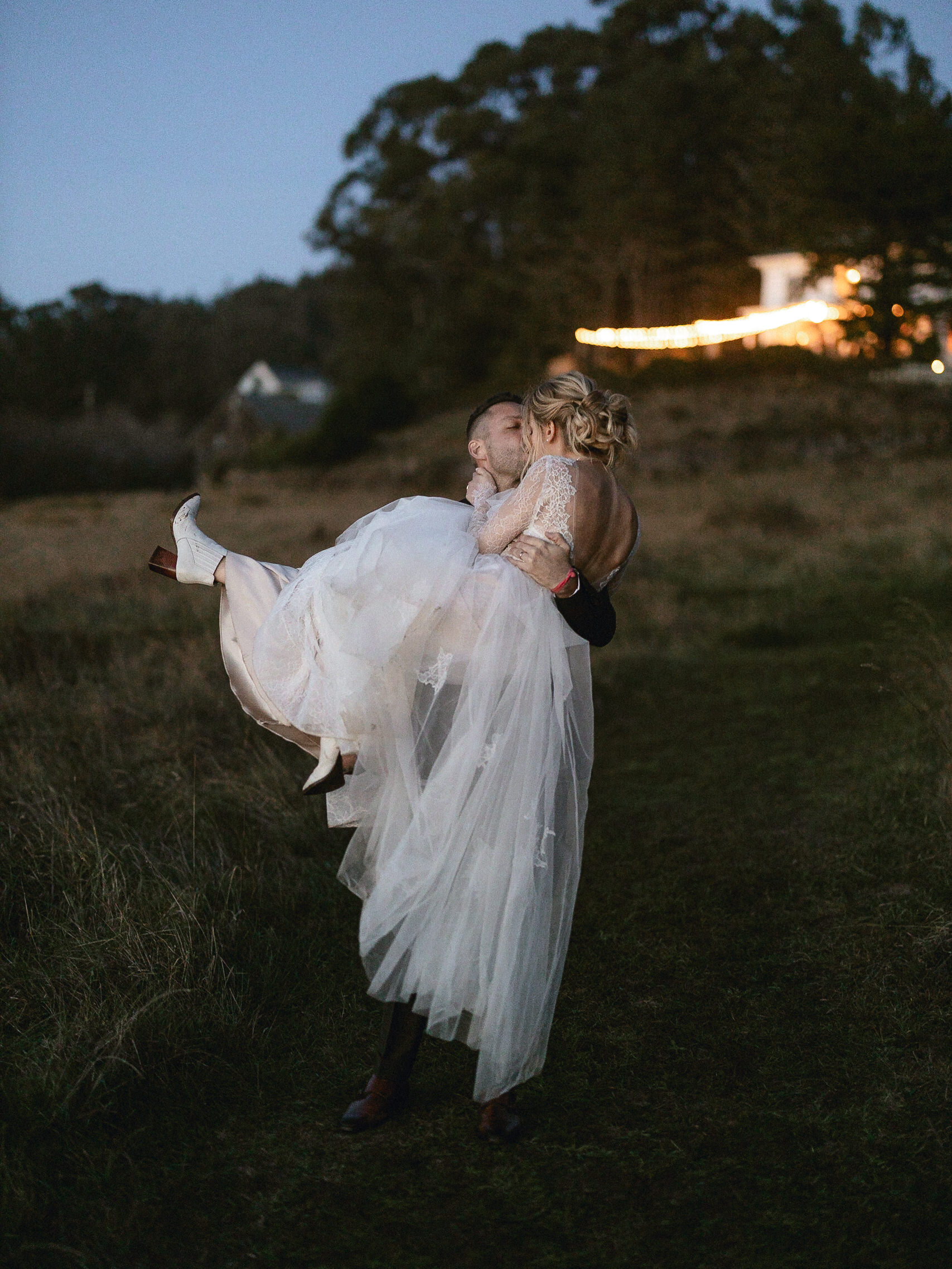 Groom carries bride in long white dress down a grassy path at dusk near Switzer Farm, with trees and a lit house in background.