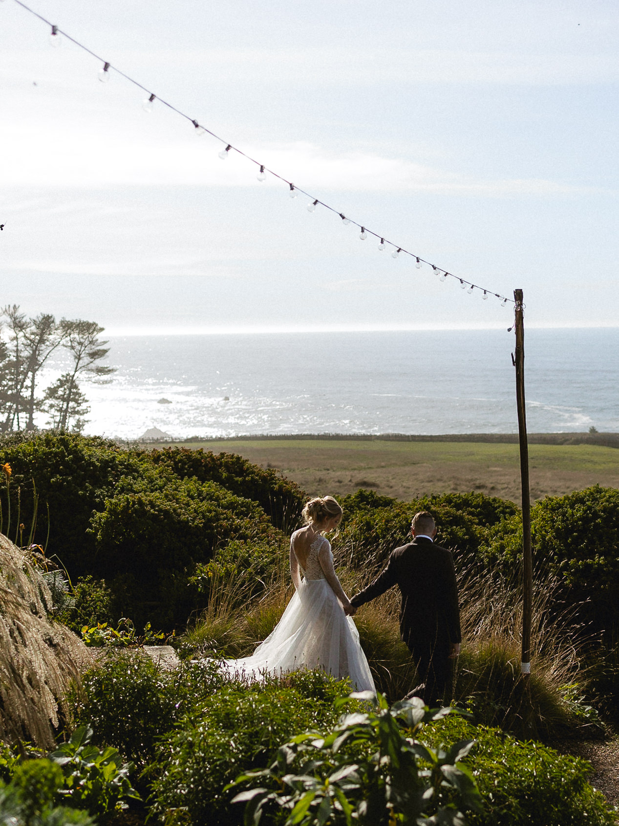 Bride and groom walk hand in hand down a greenery-lined path at Switzer Farm, with ocean views and string lights overhead.