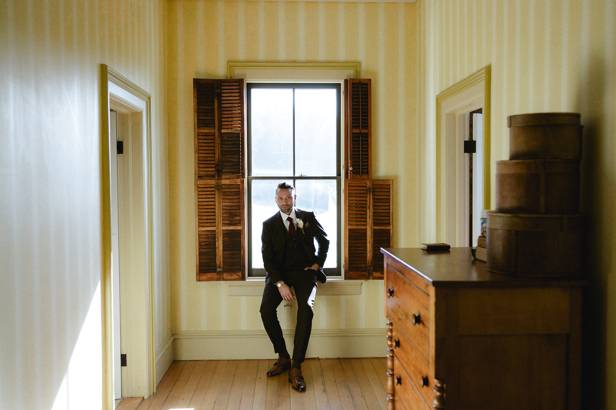 A man in a suit sits on a windowsill in a bright room with striped wallpaper, wood floors, boxes, and dresser; wedding prep.
