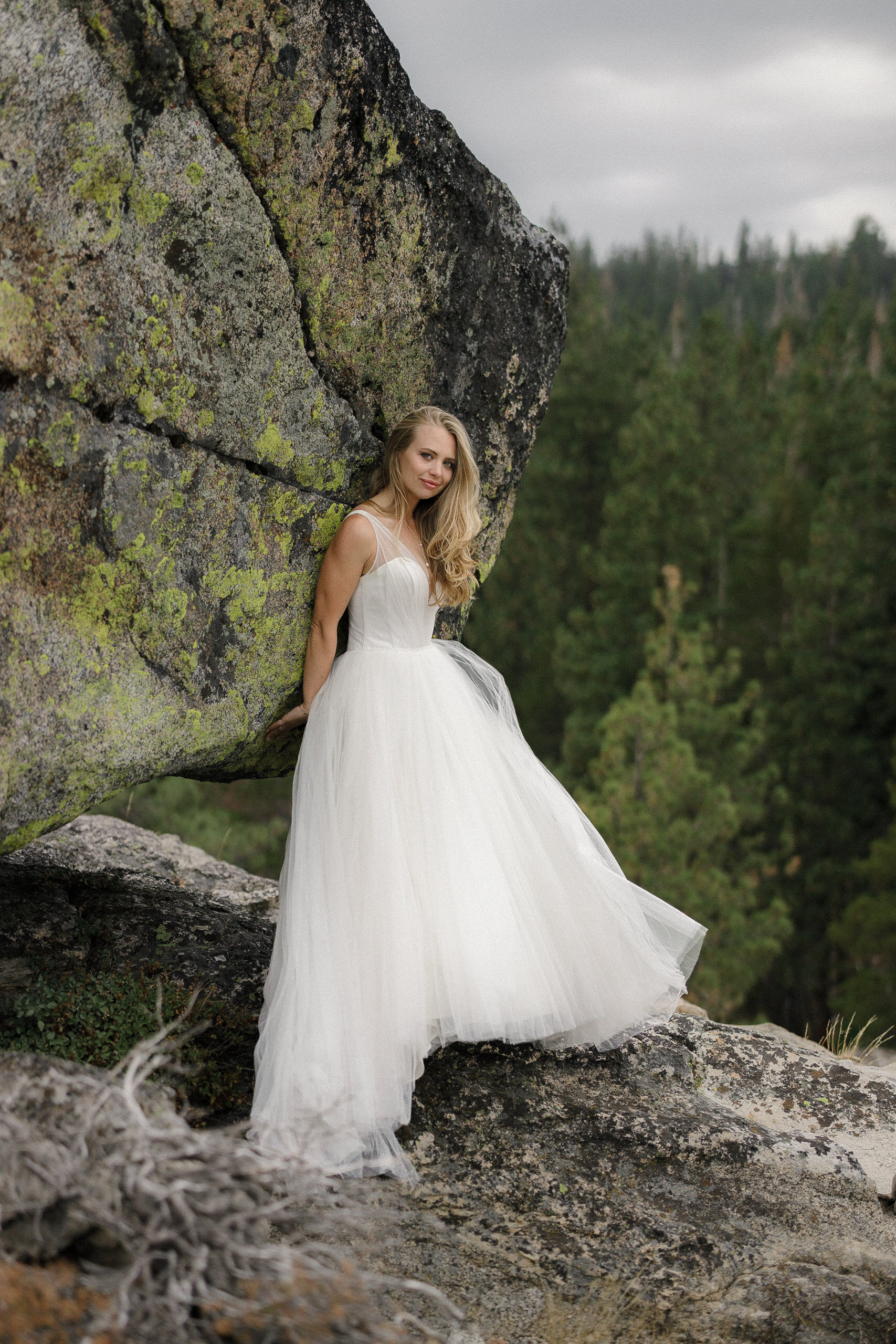 Bride in an elegant white gown poses amidst pine trees and mossy rock, showcasing serene outdoor wedding beauty.