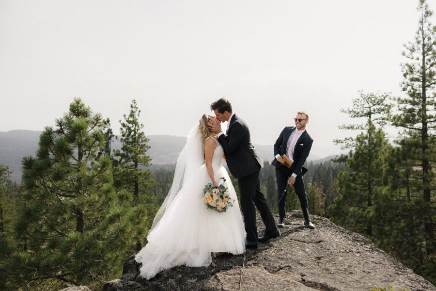 Bride and groom kiss among pine trees and mountains, captured in a stunning wedding moment.